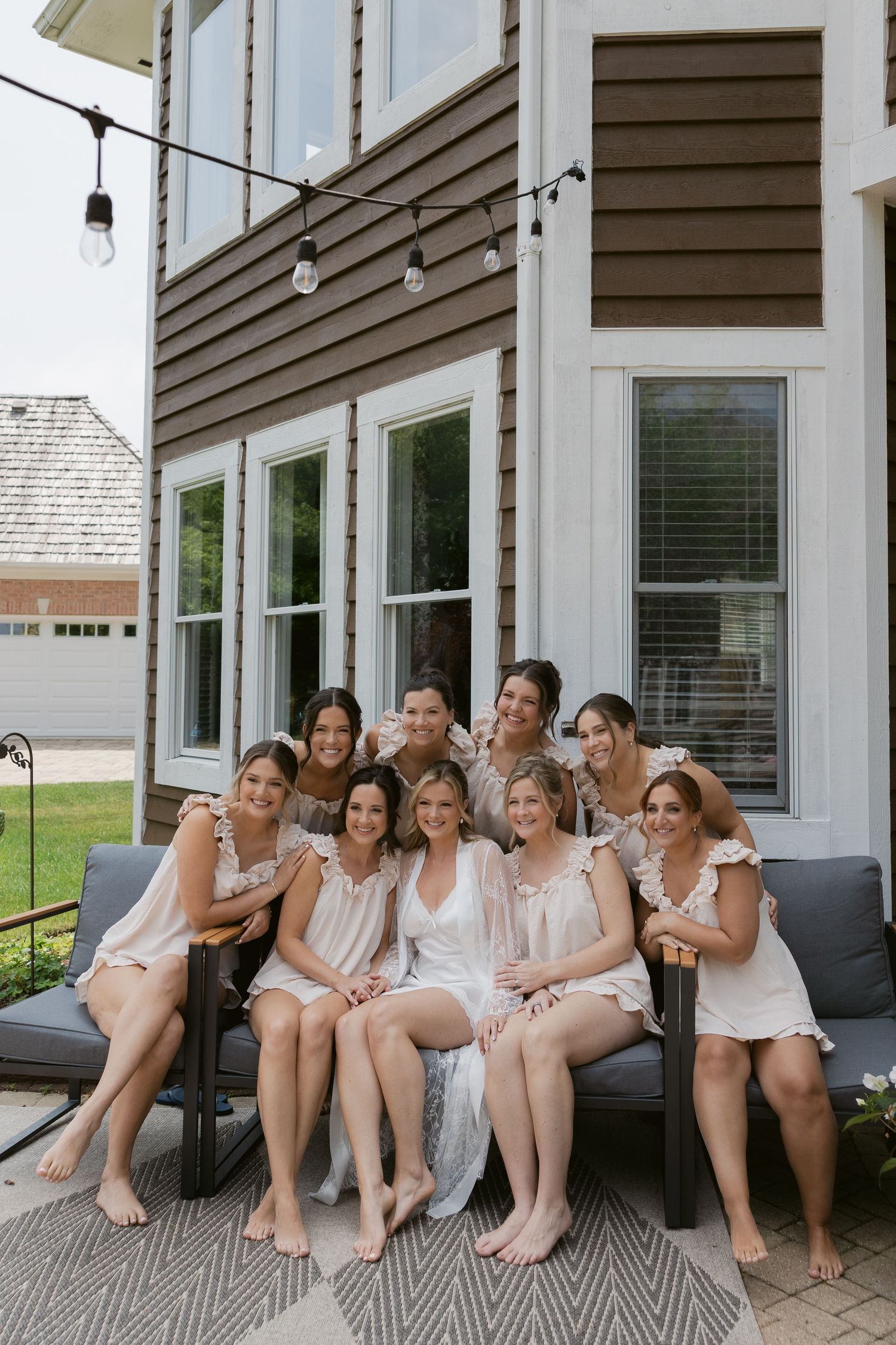 A group wearing light-colored dresses relaxes together on a covered porch with white posts and gray siding.