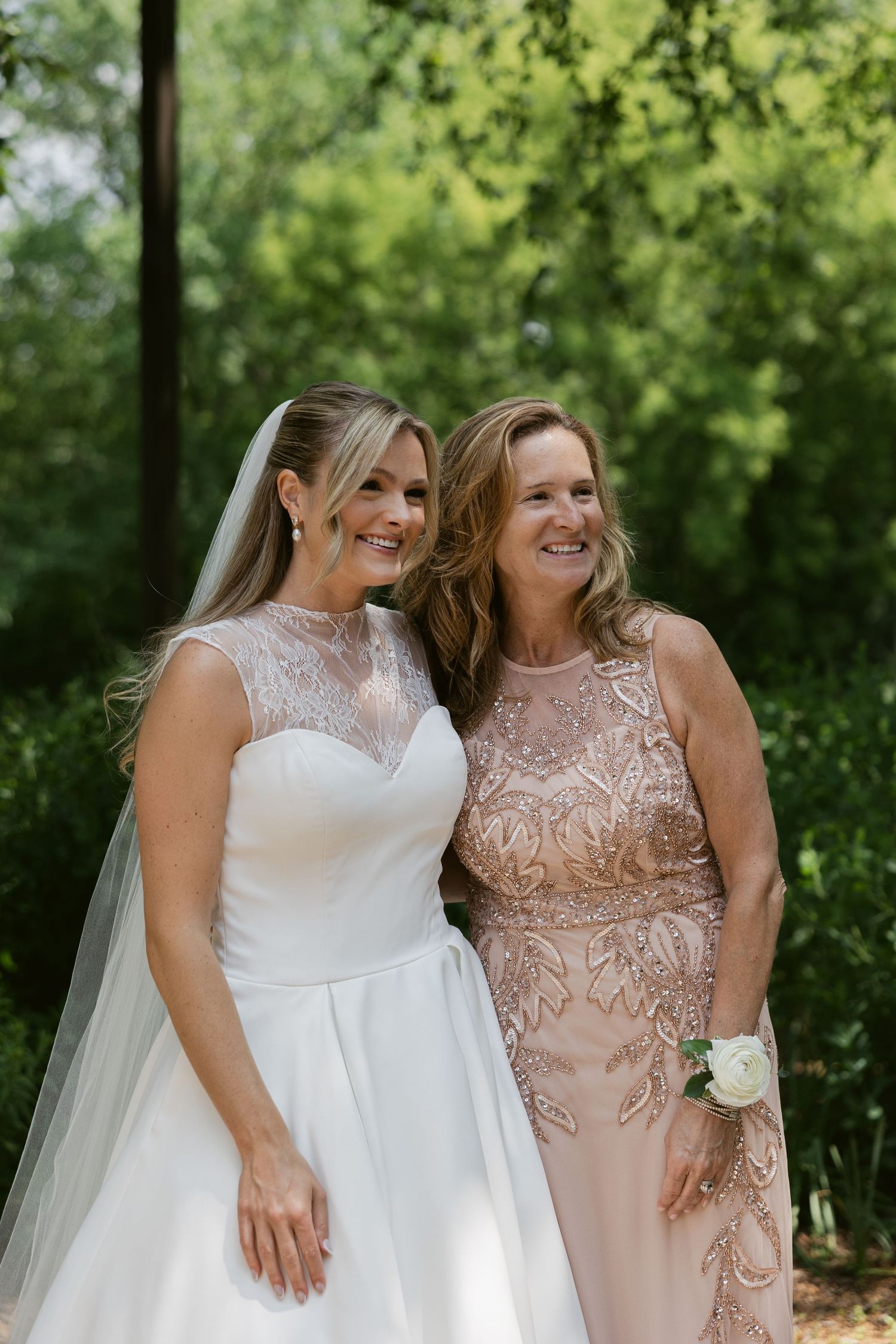 Two women share a joyful moment in a garden setting, one in a white wedding gown and one in blush formal attire.