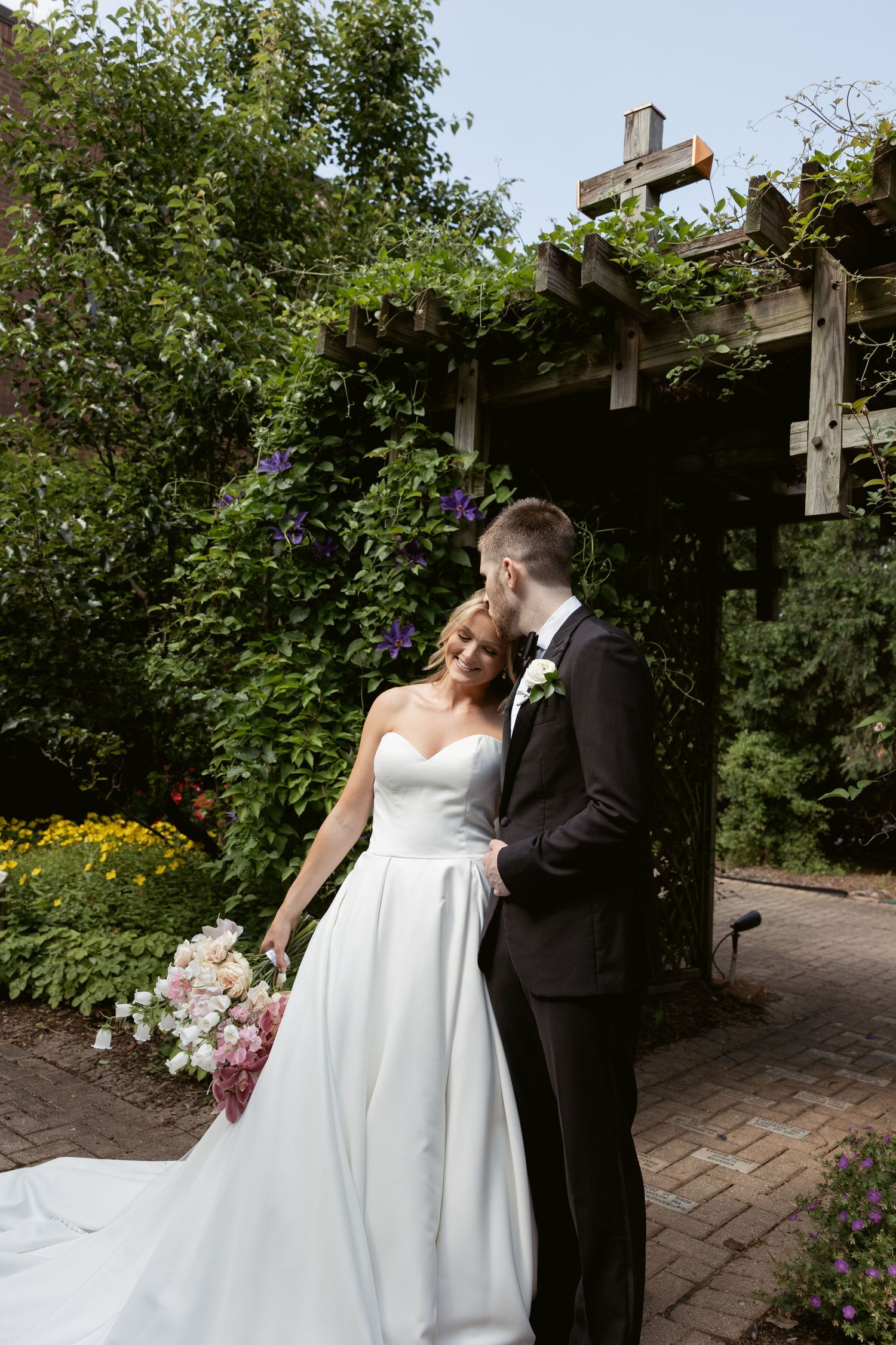 A couple embraces under a wooden pergola surrounded by blooming garden flowers.