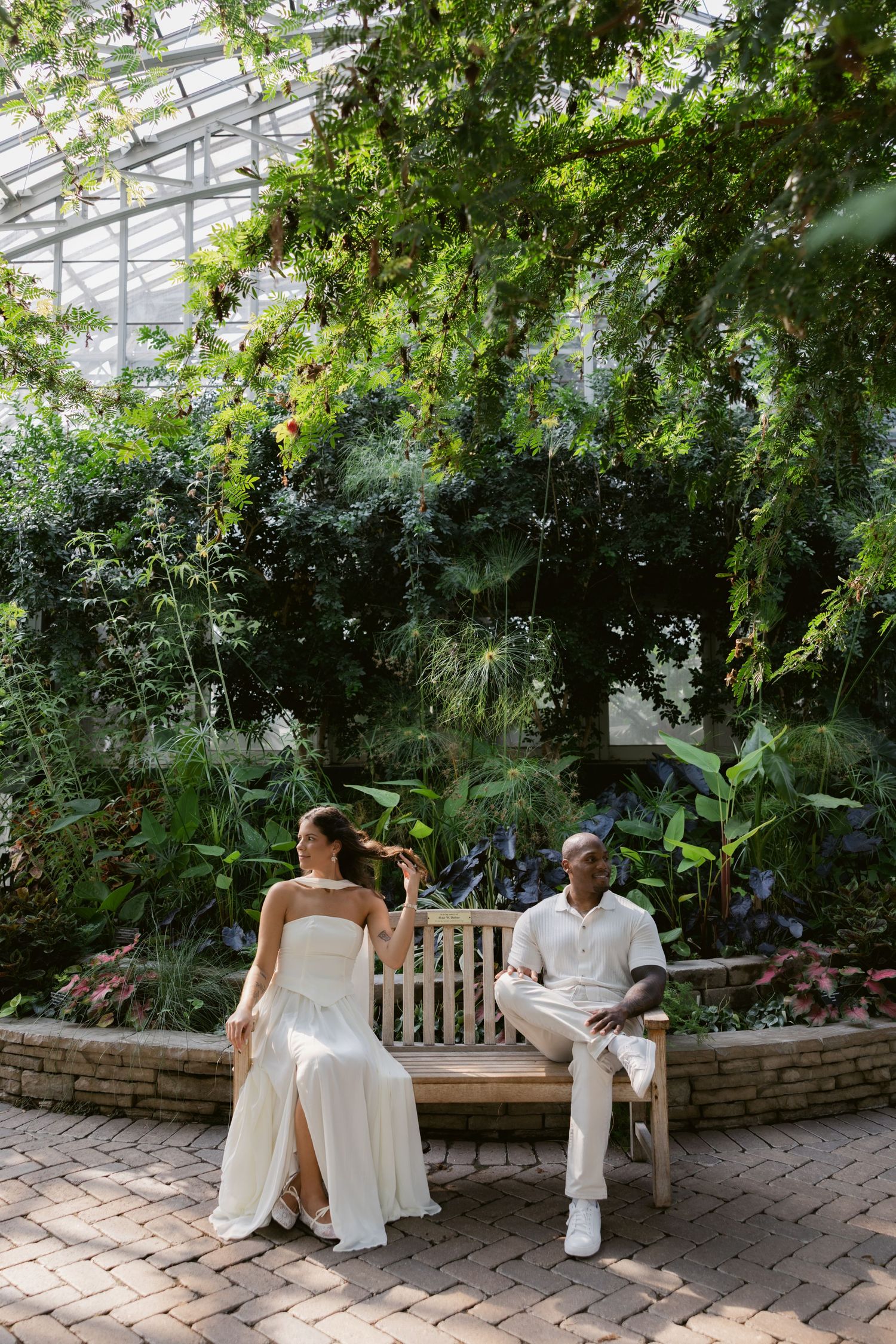 Two people in white outfits sit on a wooden bench in a lush greenhouse conservatory.