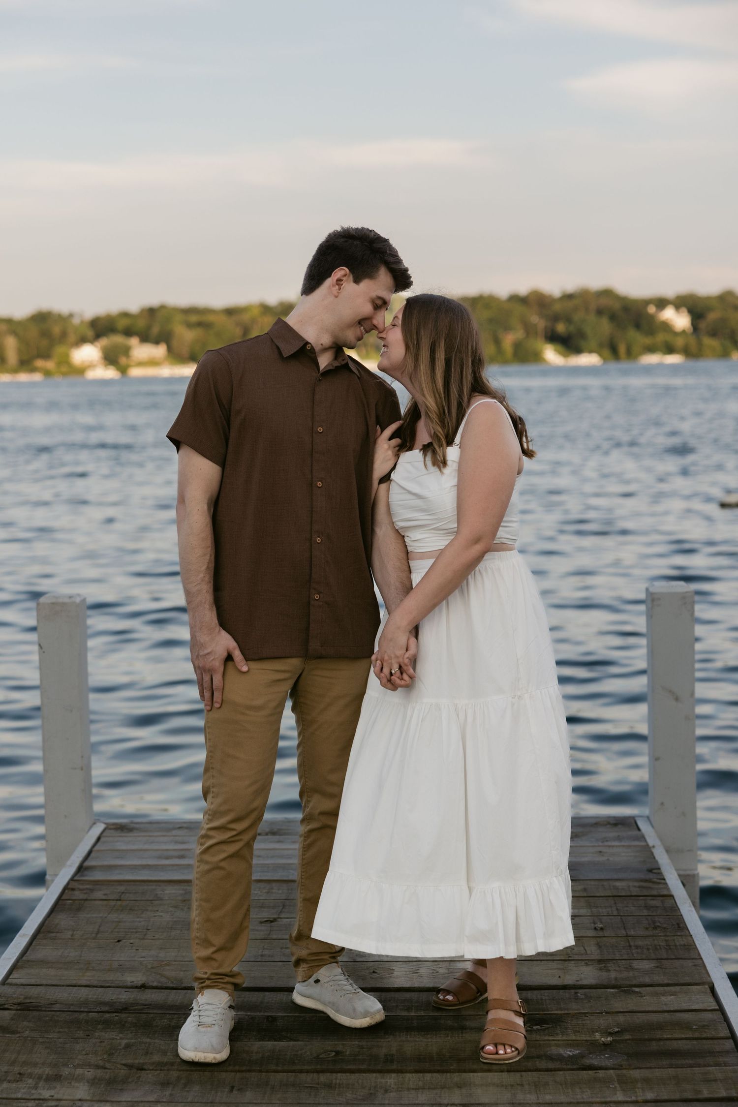 A couple stands together on a wooden dock at sunset overlooking a calm lake, dressed in casual summer attire.