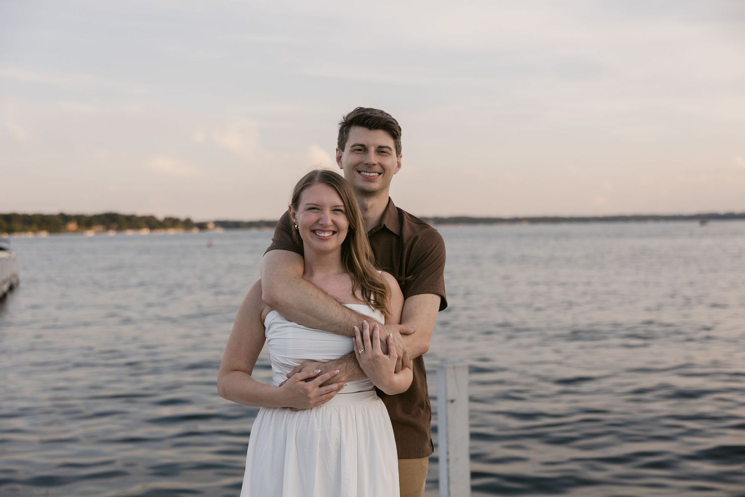A couple embraces while standing on a wooden dock with a serene lake and sunset sky in the background.