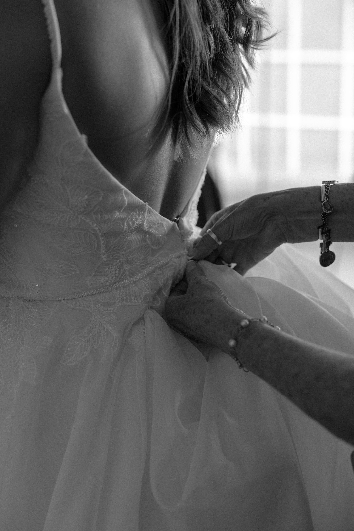 A detailed black and white photograph shows hands delicately fastening a white wedding dress in soft natural light.