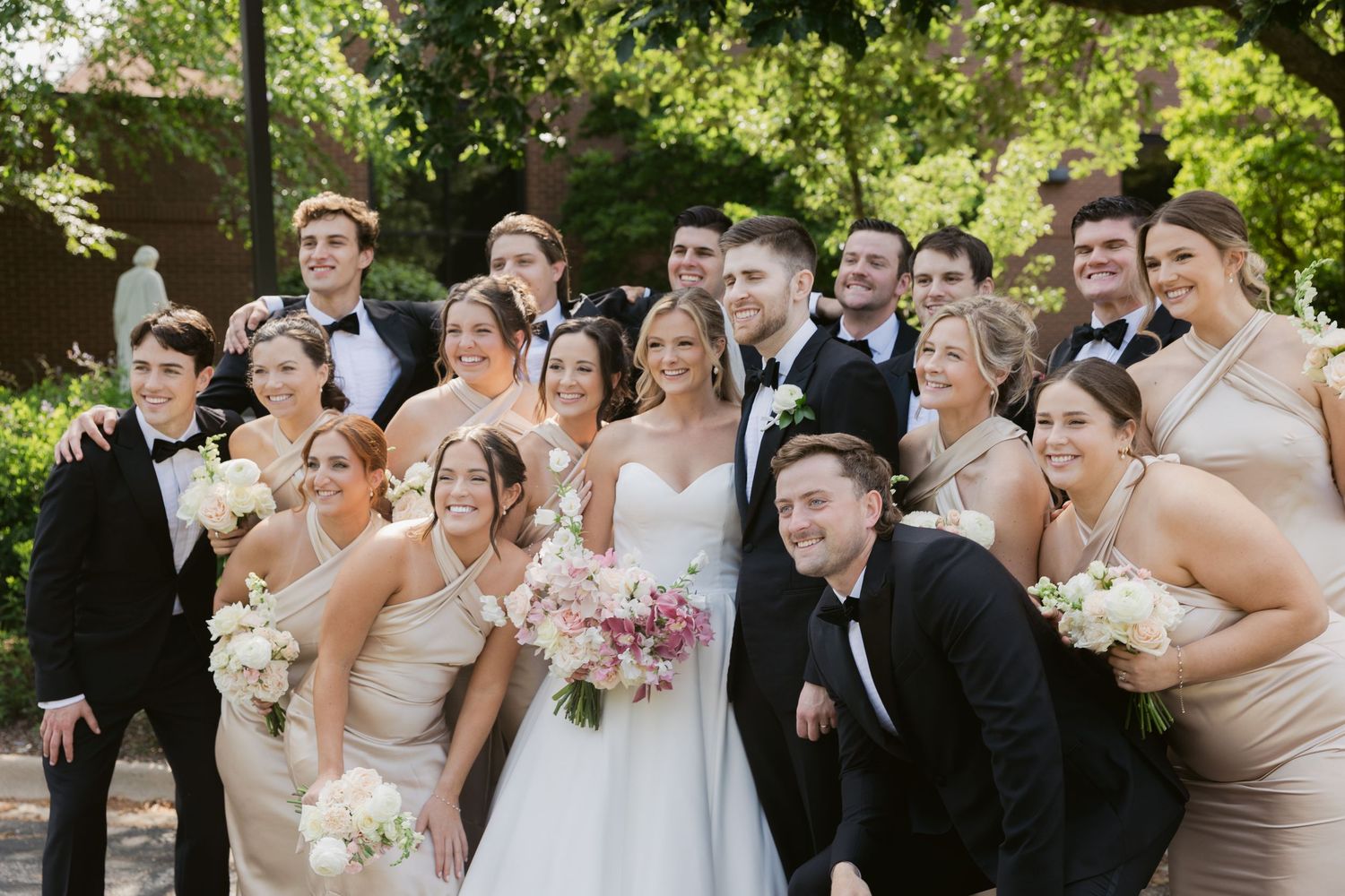 Wedding party poses together outdoors with bridesmaids in beige dresses and groomsmen in black tuxedos.