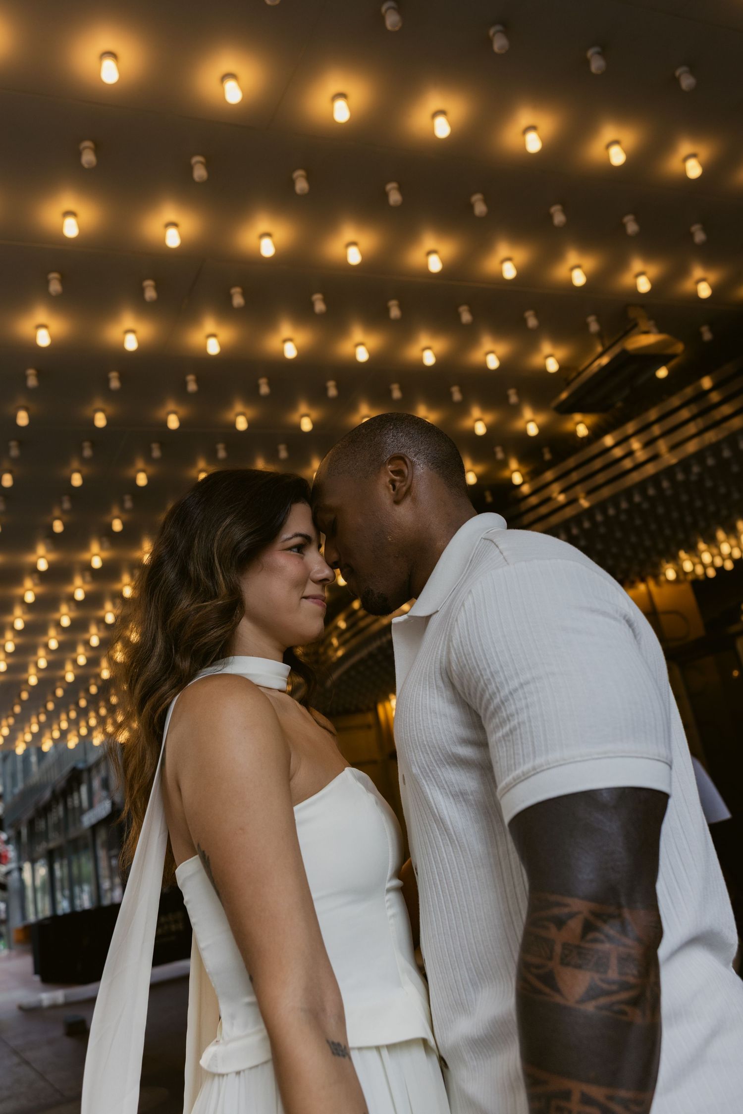 Two people in white outfits share an intimate moment under warmly lit ceiling lights.
