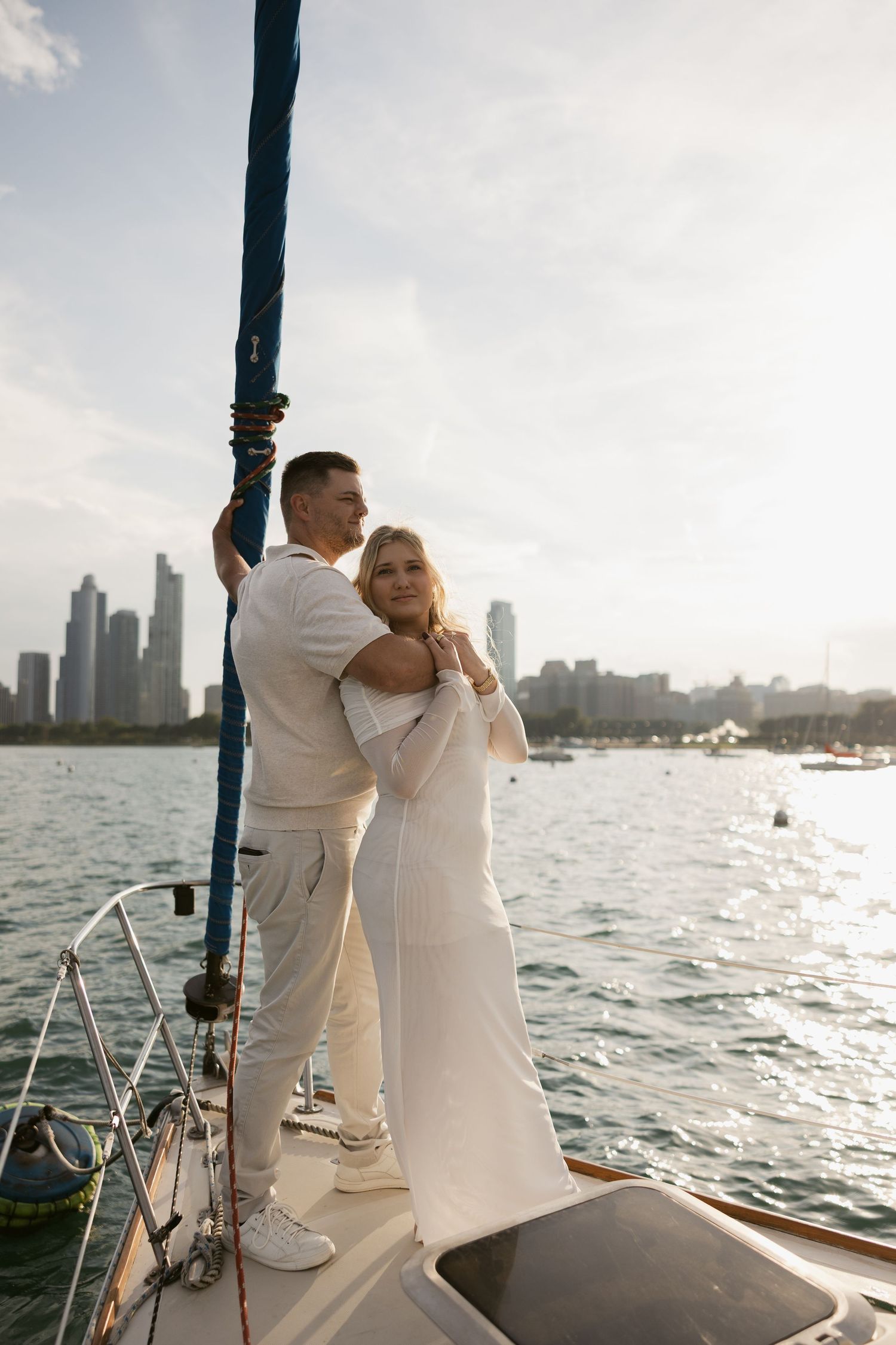 A couple in all white attire sharing a romantic moment on a sailboat with a city skyline in the background.