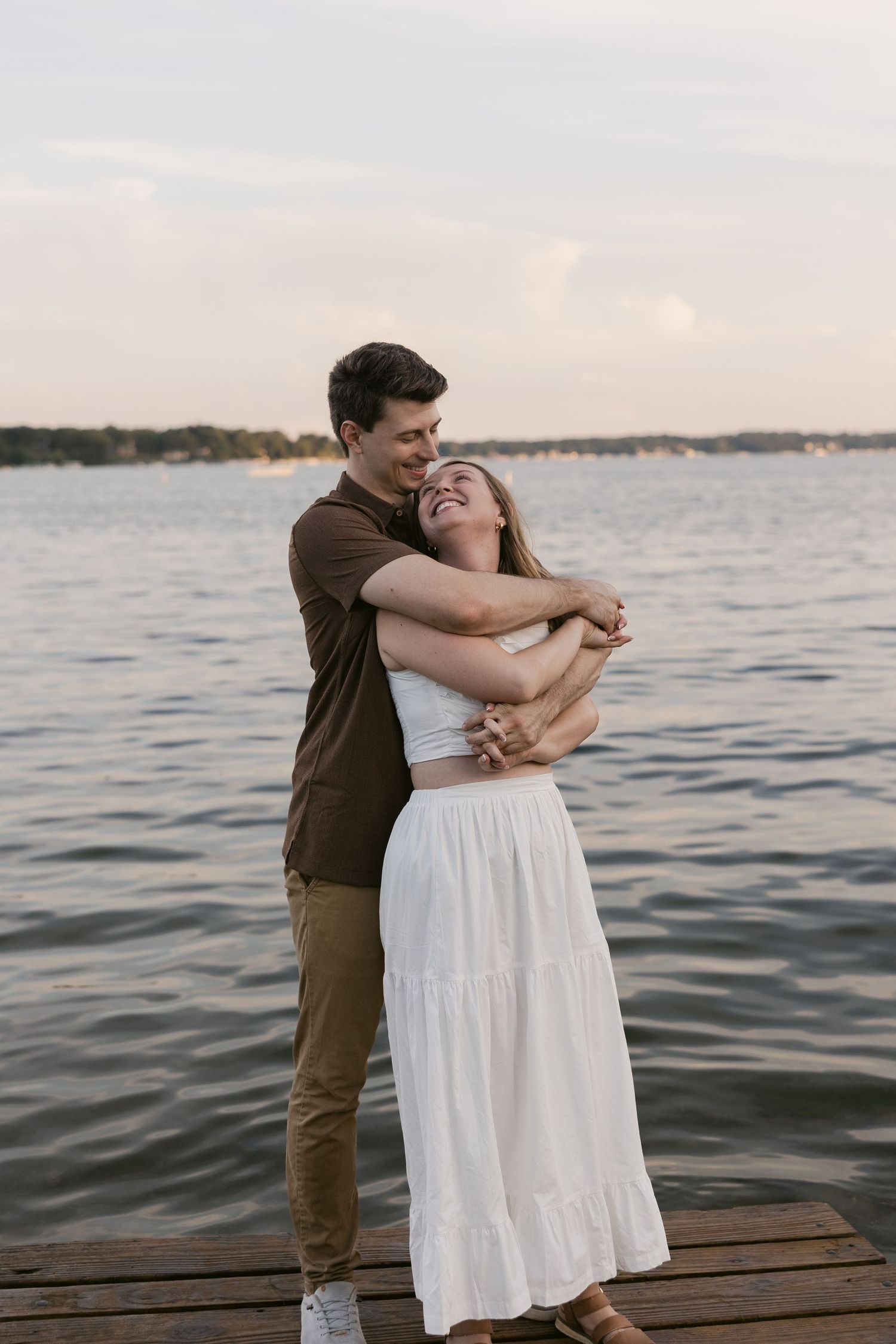 A romantic embrace between a couple standing on a wooden dock at sunset beside calm waters.