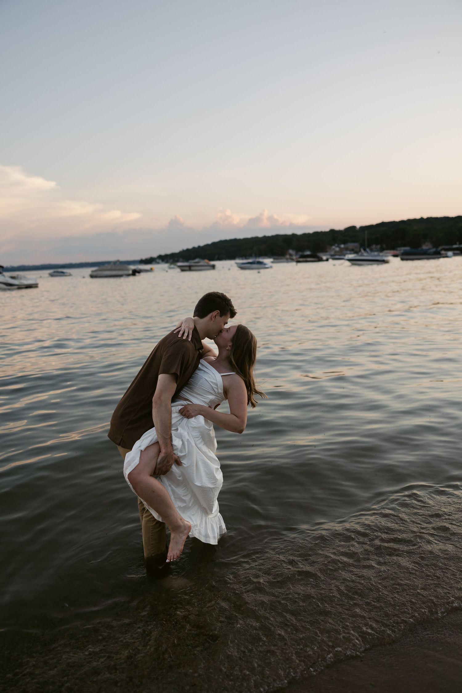 Couple plays in shallow water at sunset with boats anchored in distance.