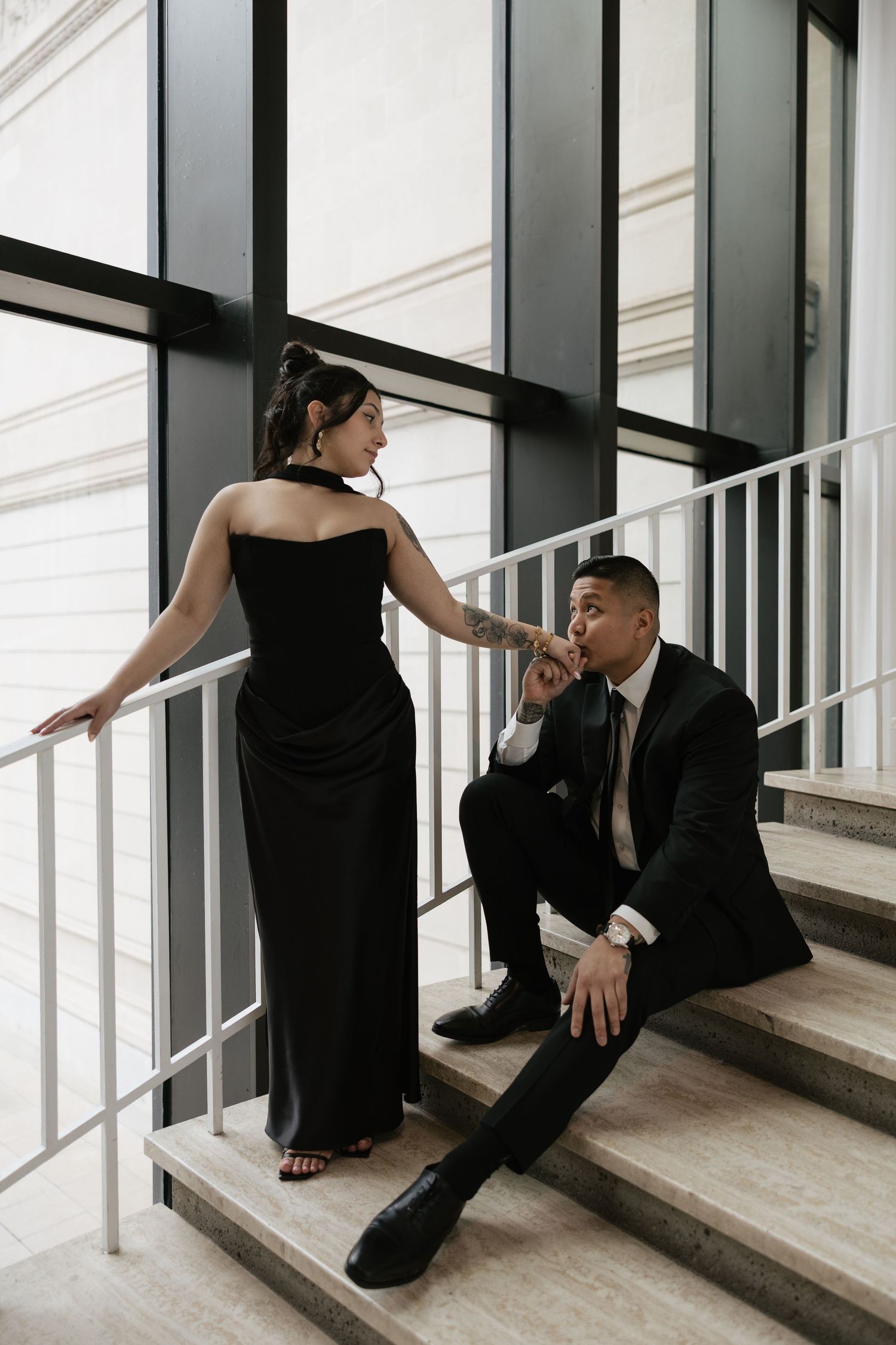 A couple in black attire poses intimately on concrete stairs in an artistic photoshoot series.