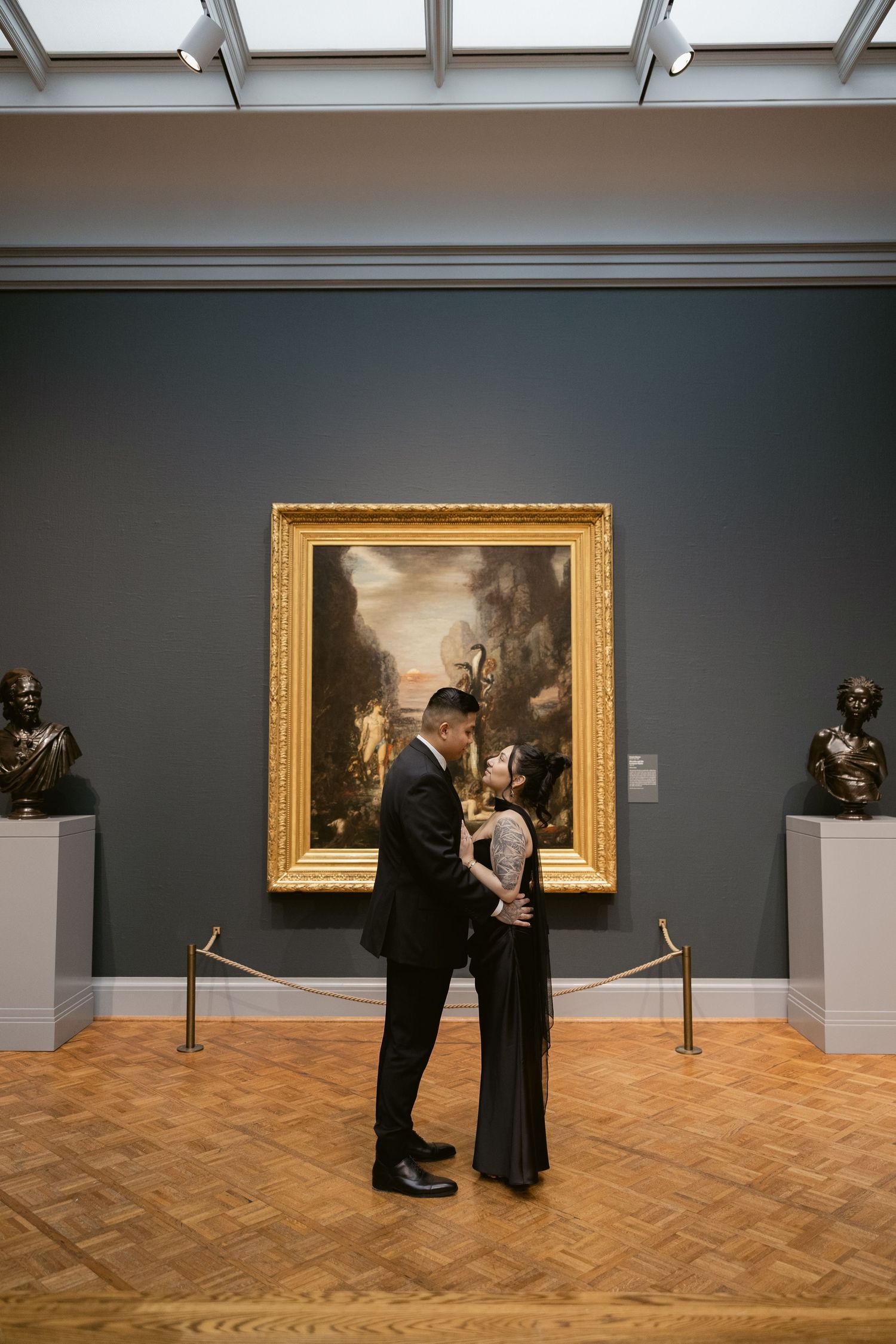 A couple views artwork in an ornate gold frame at an art museum gallery with parquet flooring.