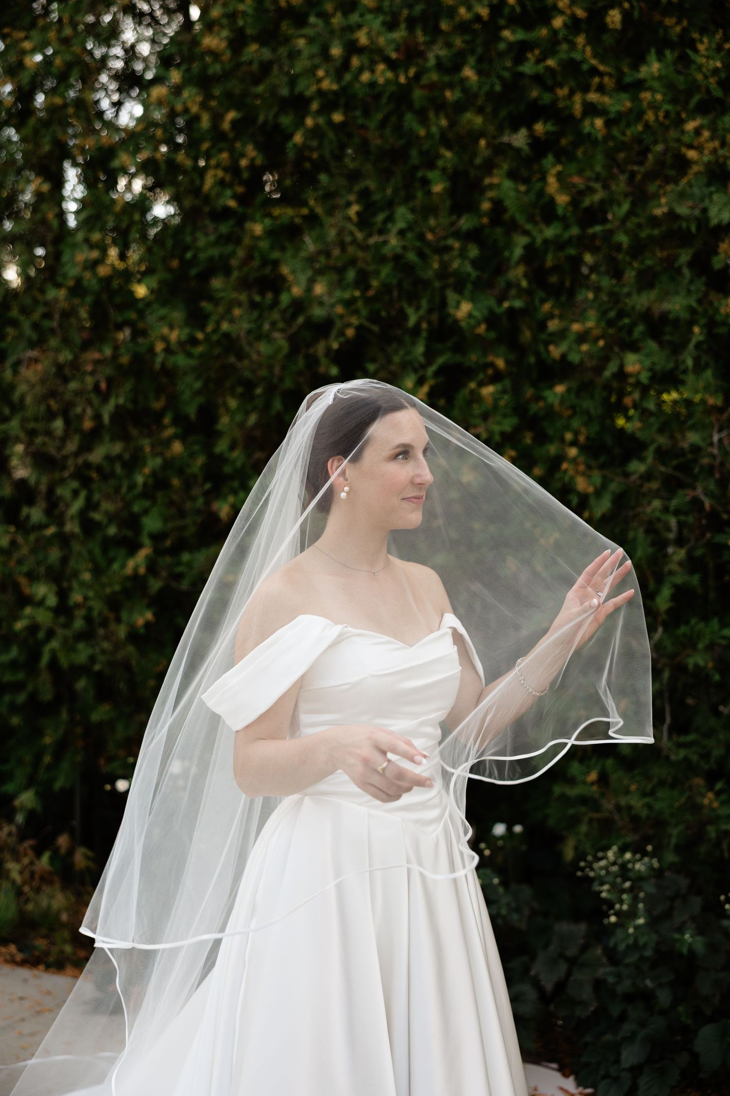 A bride in a flowing white wedding dress and veil poses gracefully against a lush green garden backdrop.