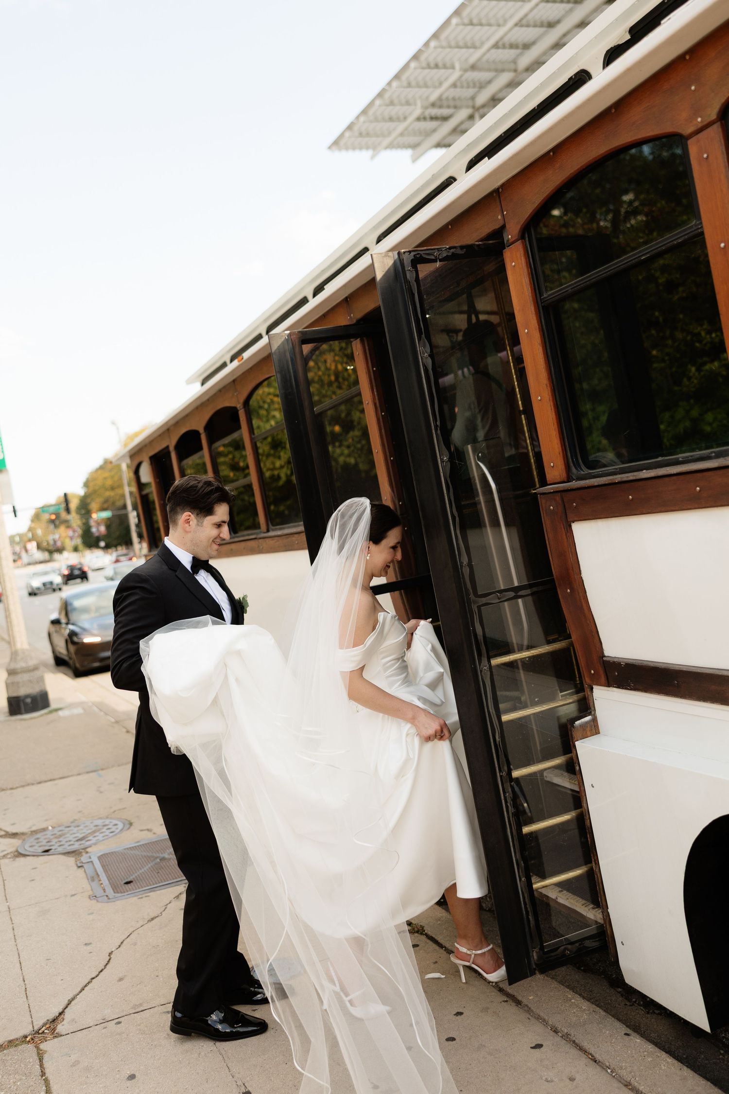 A couple in formal wedding attire board a vintage trolley car with flowing white dress trailing behind them.