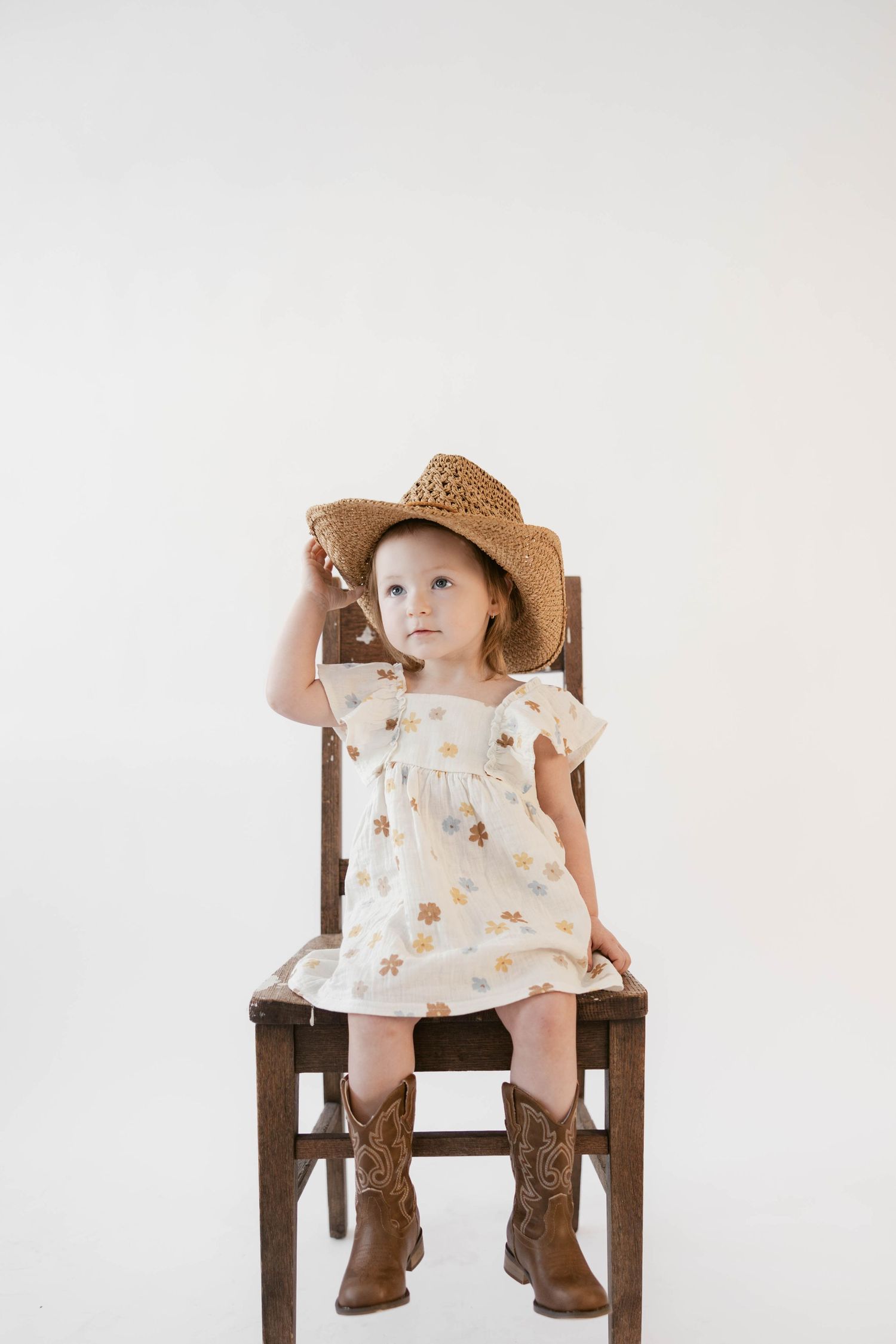 Child in floral dress and straw hat poses on wooden chair during photo session.