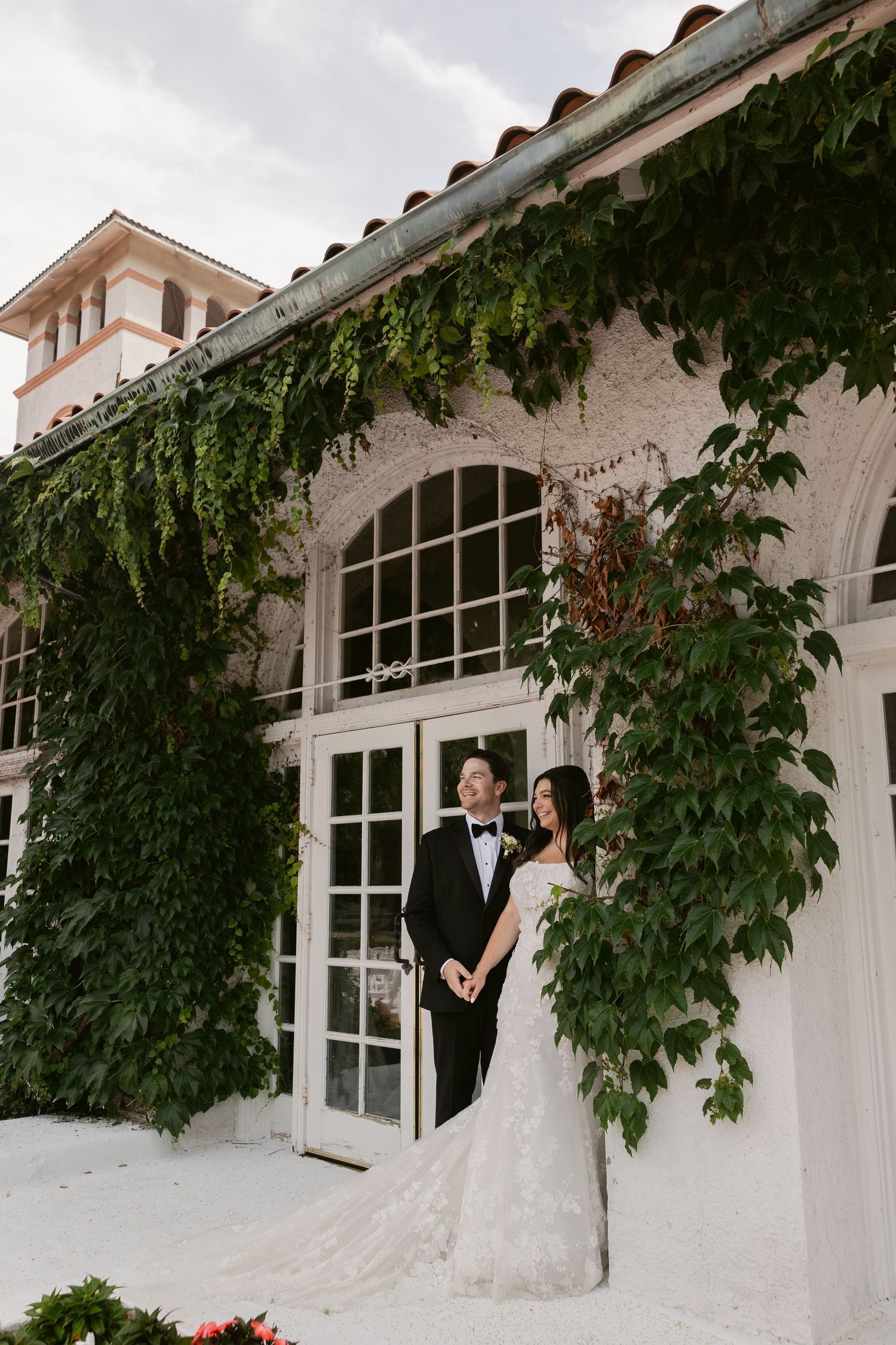 A wedding couple stands in front of a white Spanish-style building with ivy-covered walls and arched windows.