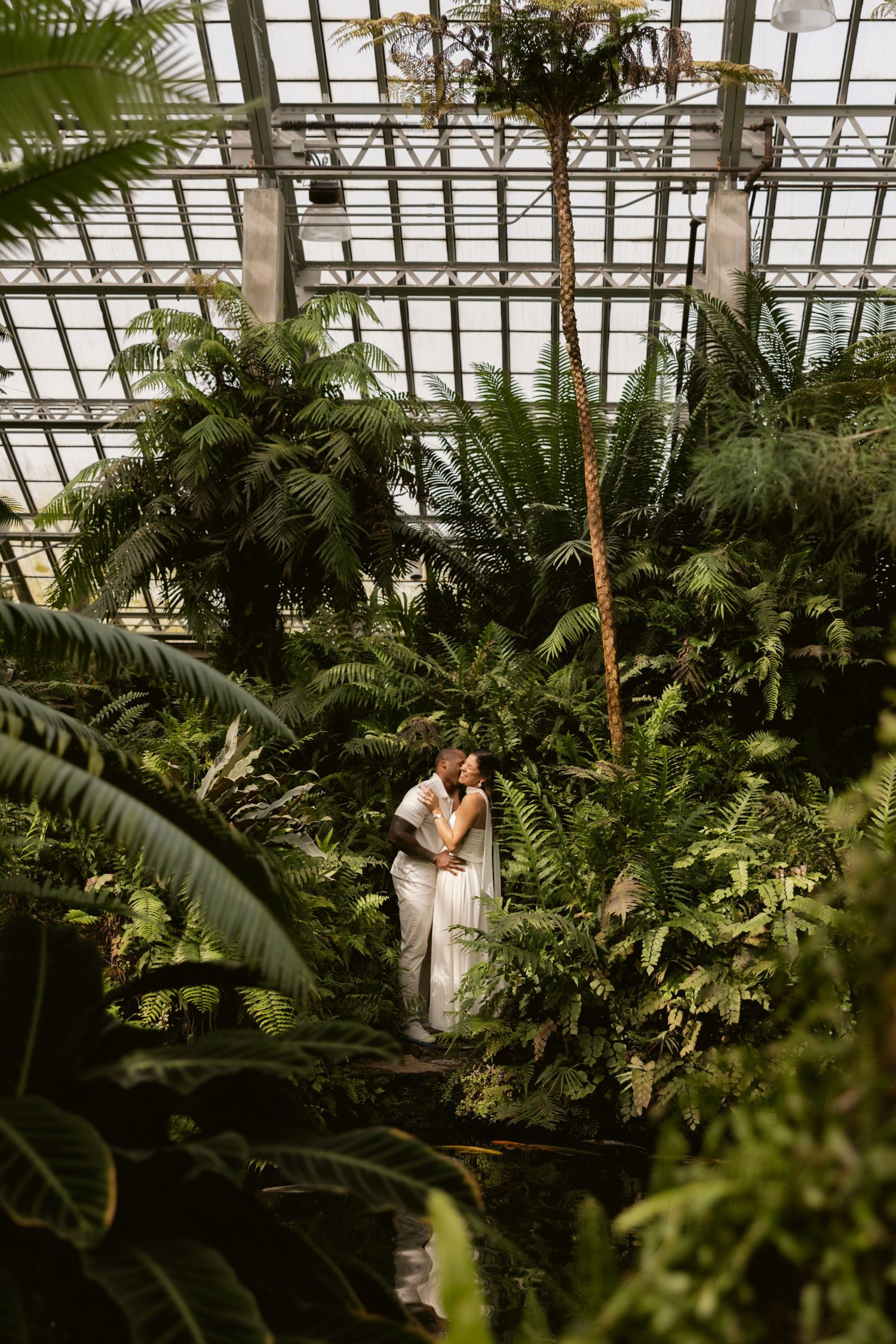 A romantic greenhouse wedding photoshoot among dense tropical foliage and glass ceiling architecture.
