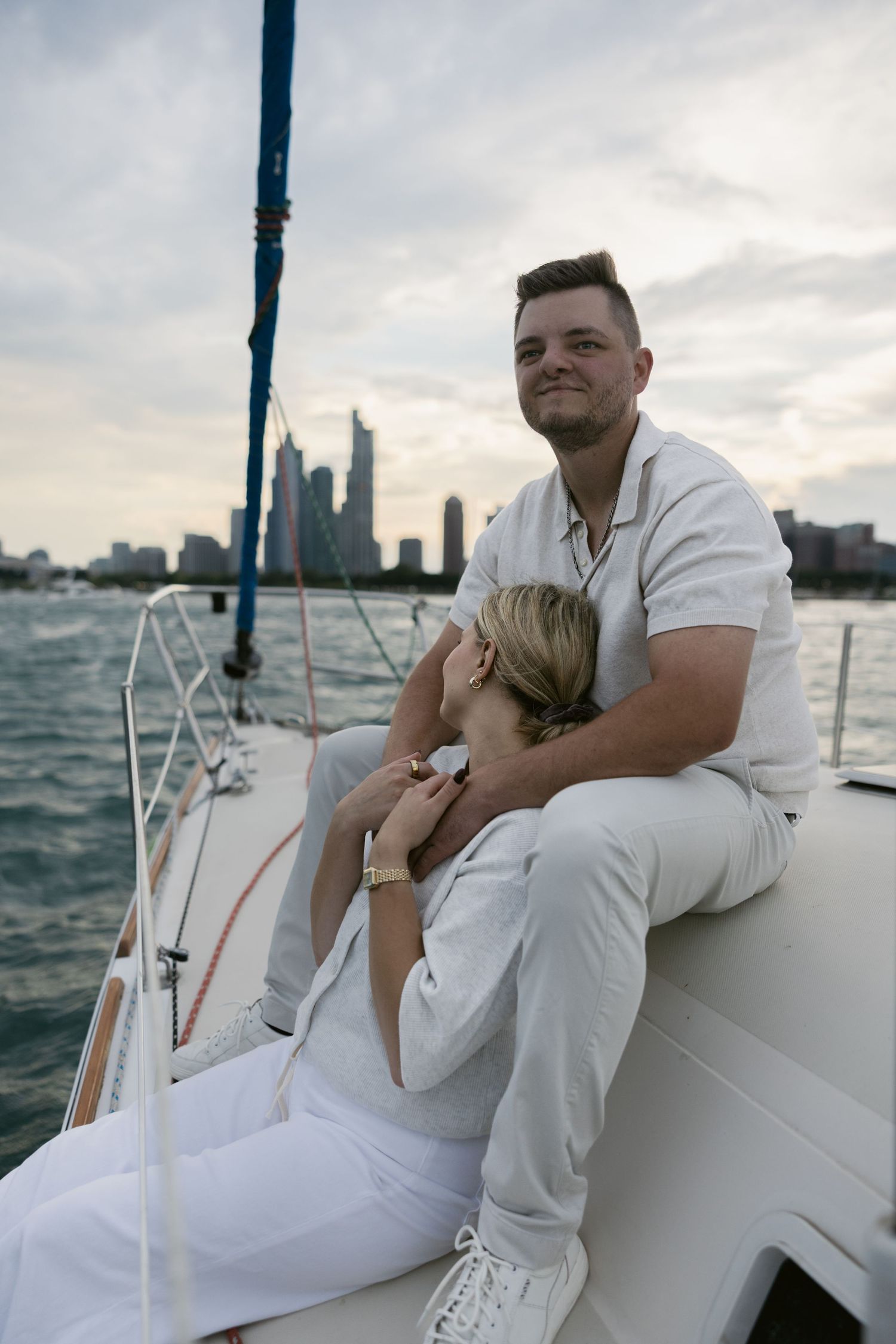 A couple in white clothing sits together on a sailboat against an urban waterfront backdrop.