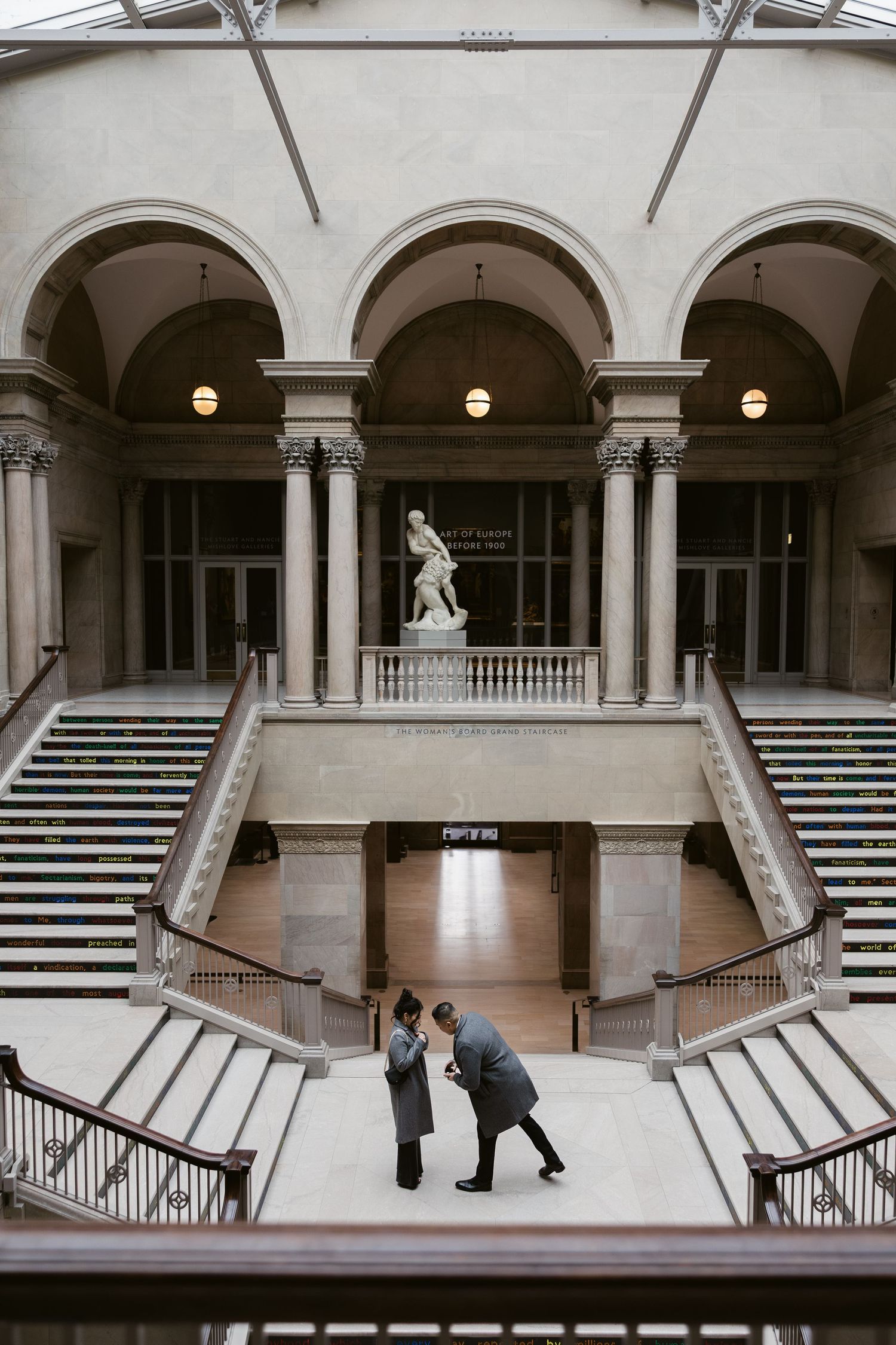 Two figures meet on symmetrical marble staircases beneath arched columns and a classical statue in a museum atrium.