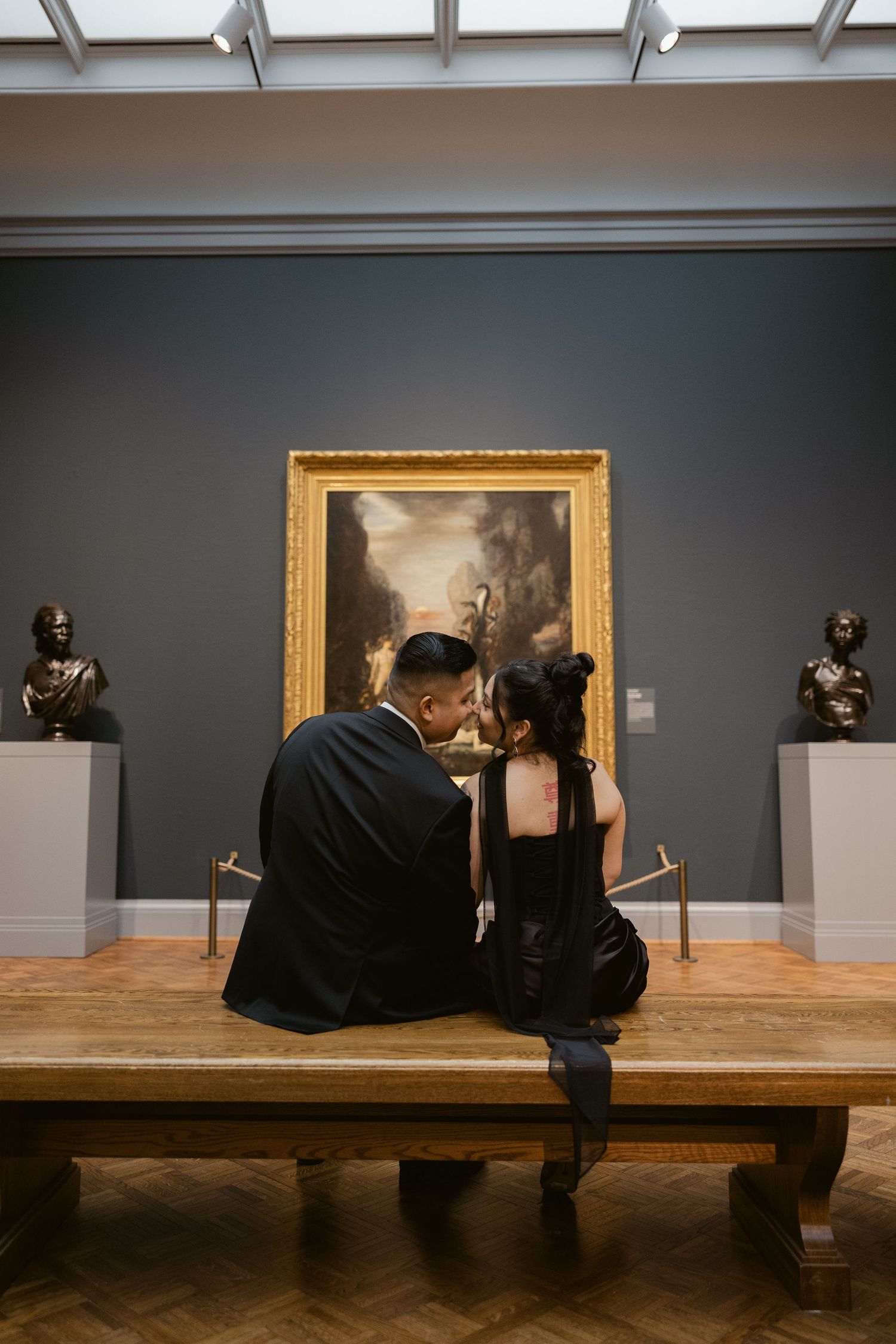 Two people in dark clothing sit on a wooden bench in an art gallery surrounded by paintings.