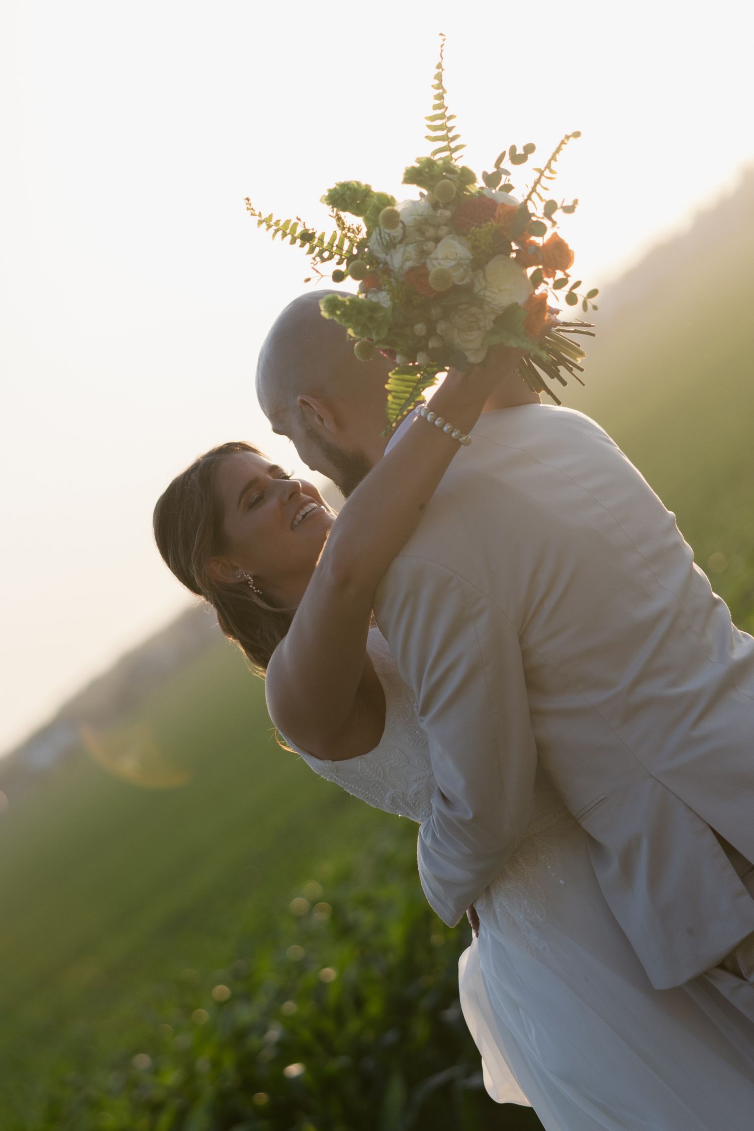 A romantic wedding couple embraces in golden sunlight while holding a delicate bouquet of ferns and flowers.