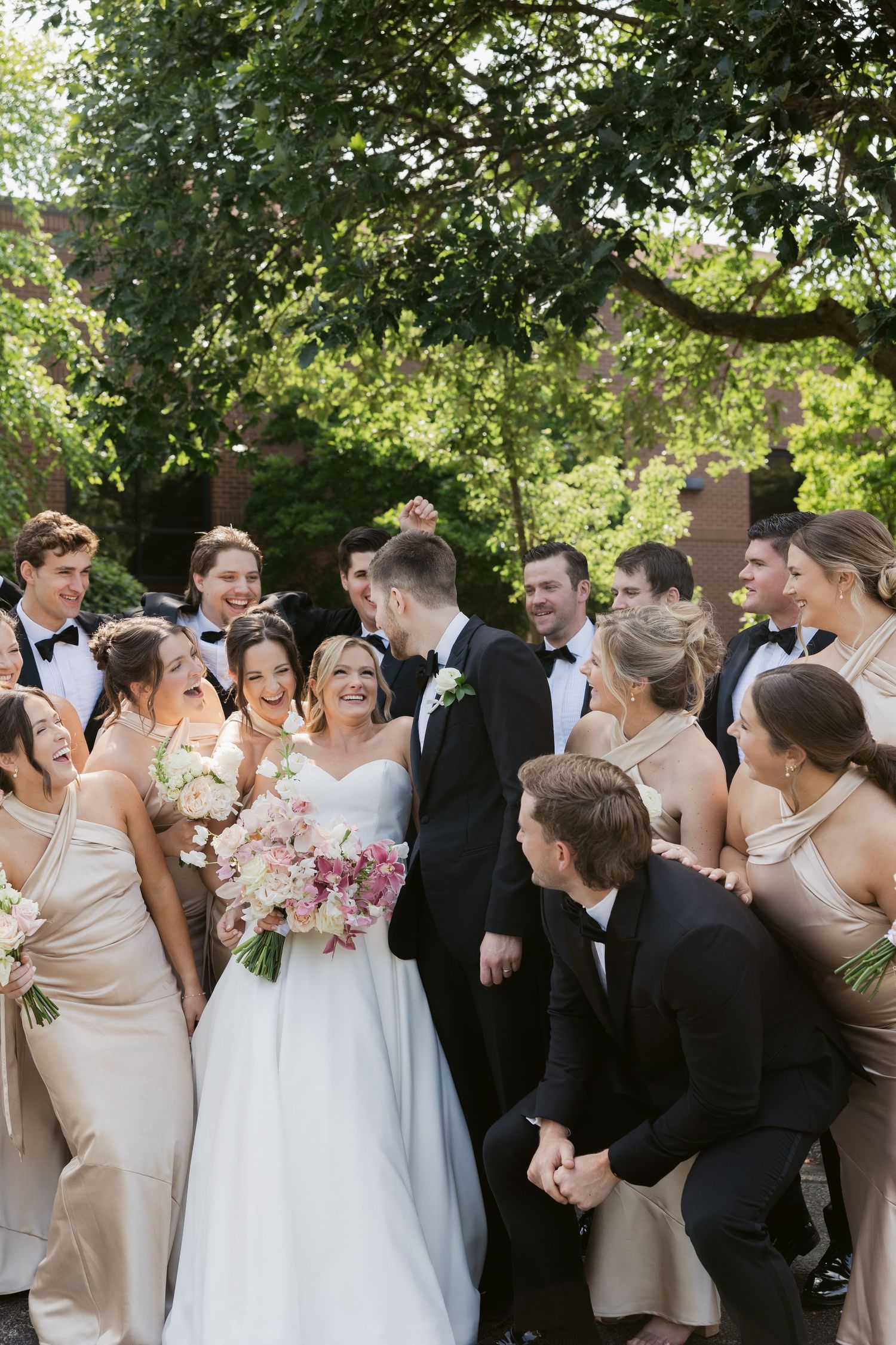 Wedding party gathered outdoors with bridesmaids in champagne dresses and groomsmen in black tuxedos.