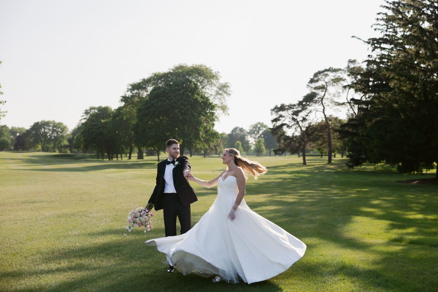 A couple dances and twirls together on a sunny golf course with lush green grass and trees in the background.