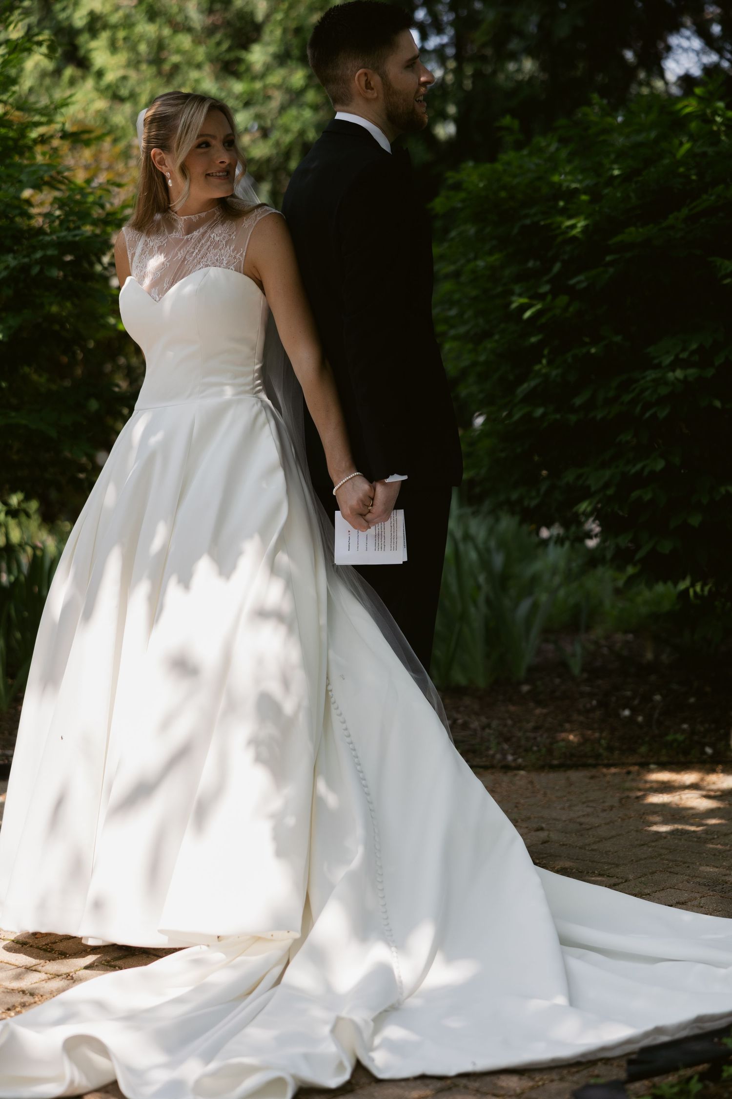 Elegant bride in flowing white gown with long train posing in outdoor garden setting.