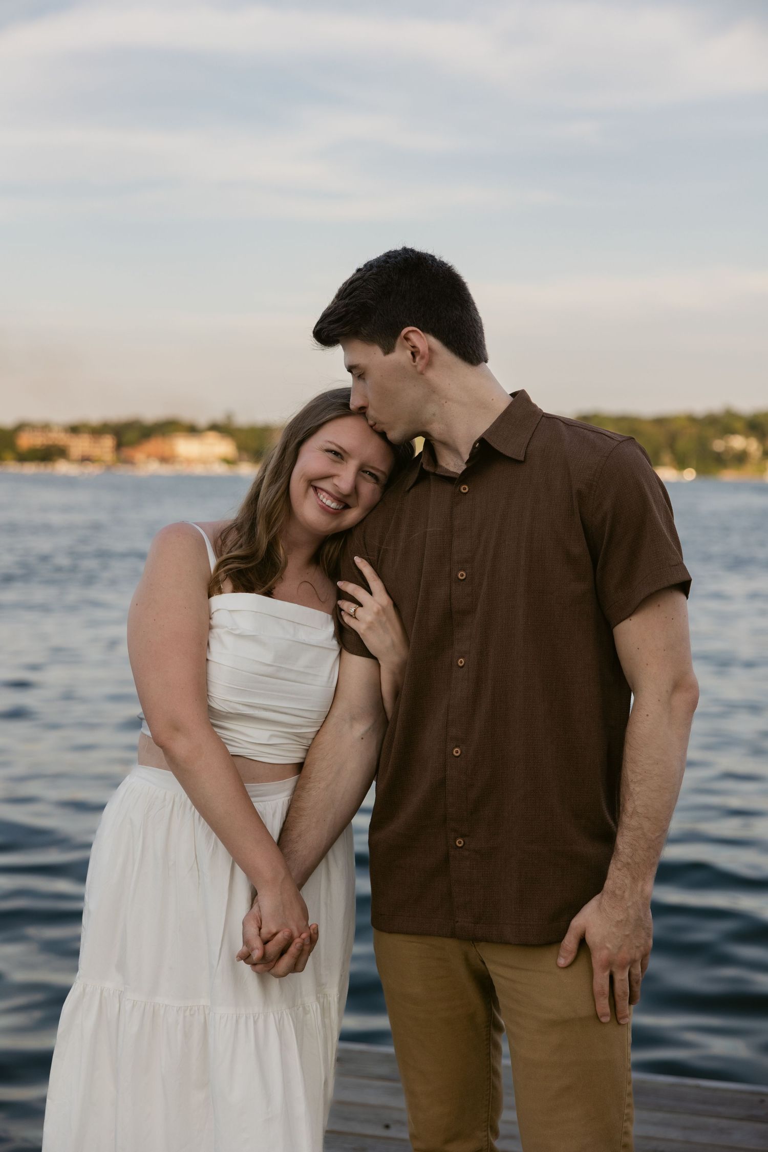 A romantic seaside photoshoot series shows a couple in white dress and brown shirt embracing by the water at sunset.
