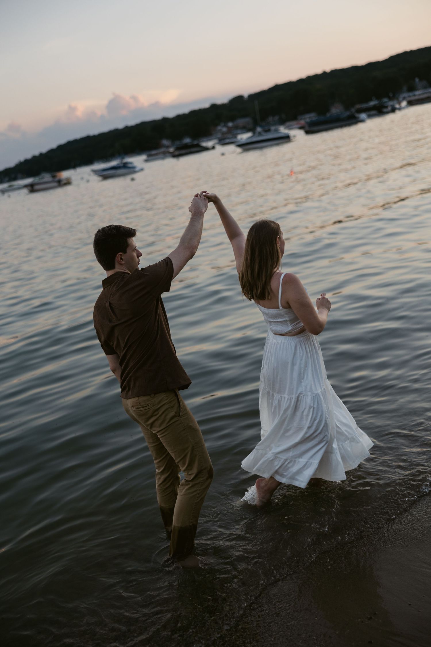 A couple walks at the water's edge during sunset as boats float in the distance.
