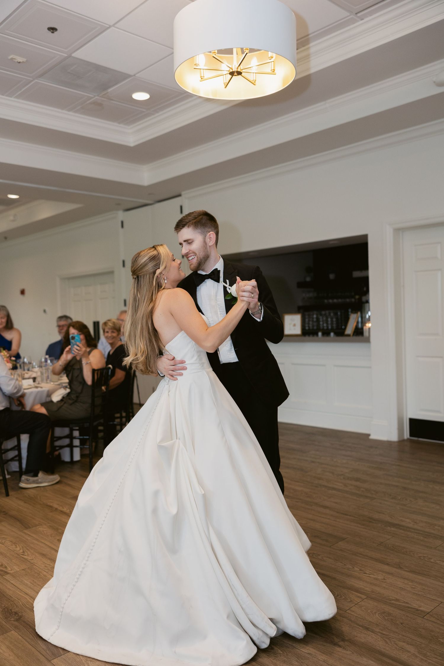 Couple performs romantic first dance together in an elegant ballroom with warm lighting overhead.