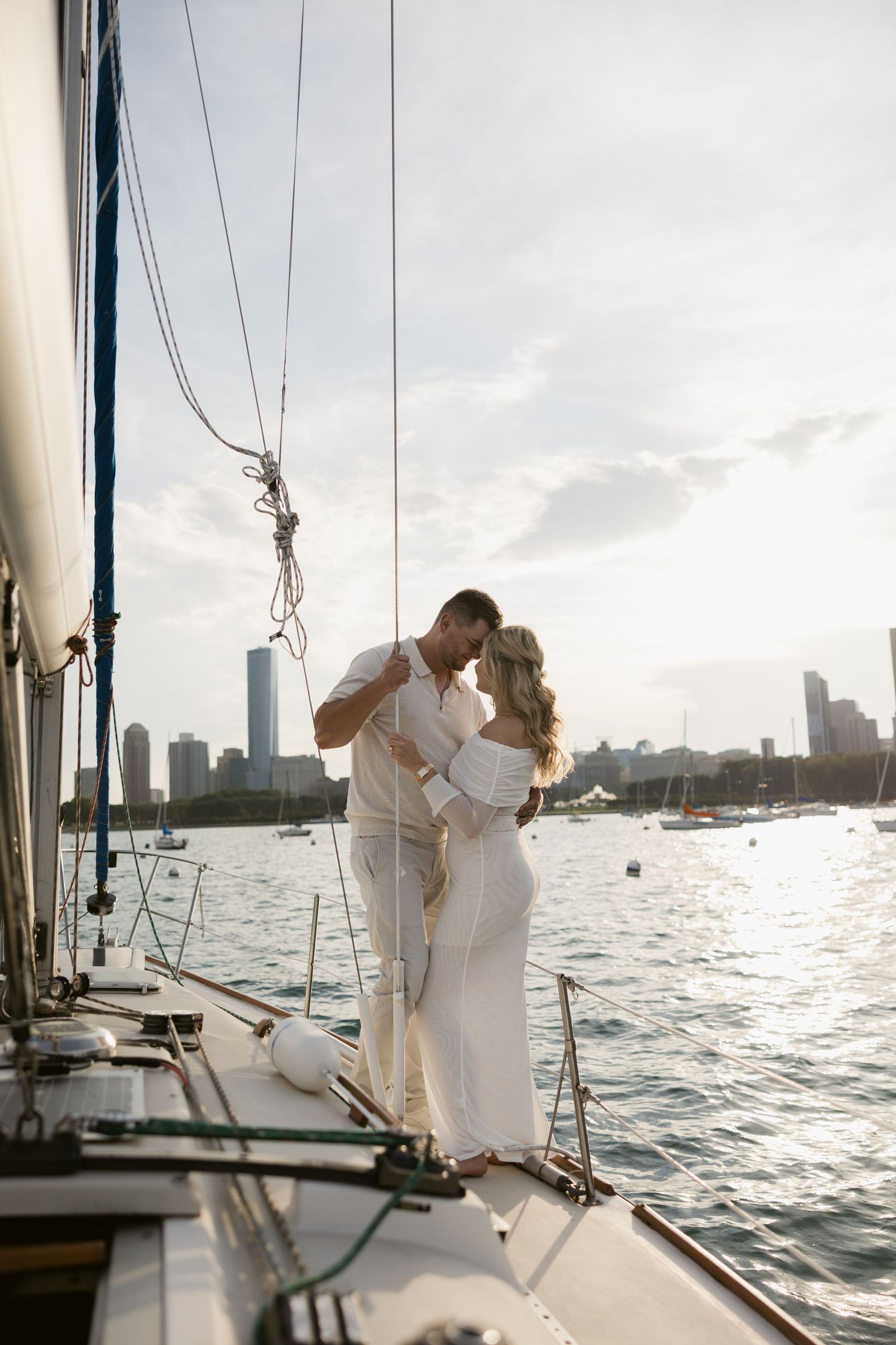 A couple dressed in white embraces on the bow of a sailboat with a city skyline backdrop.
