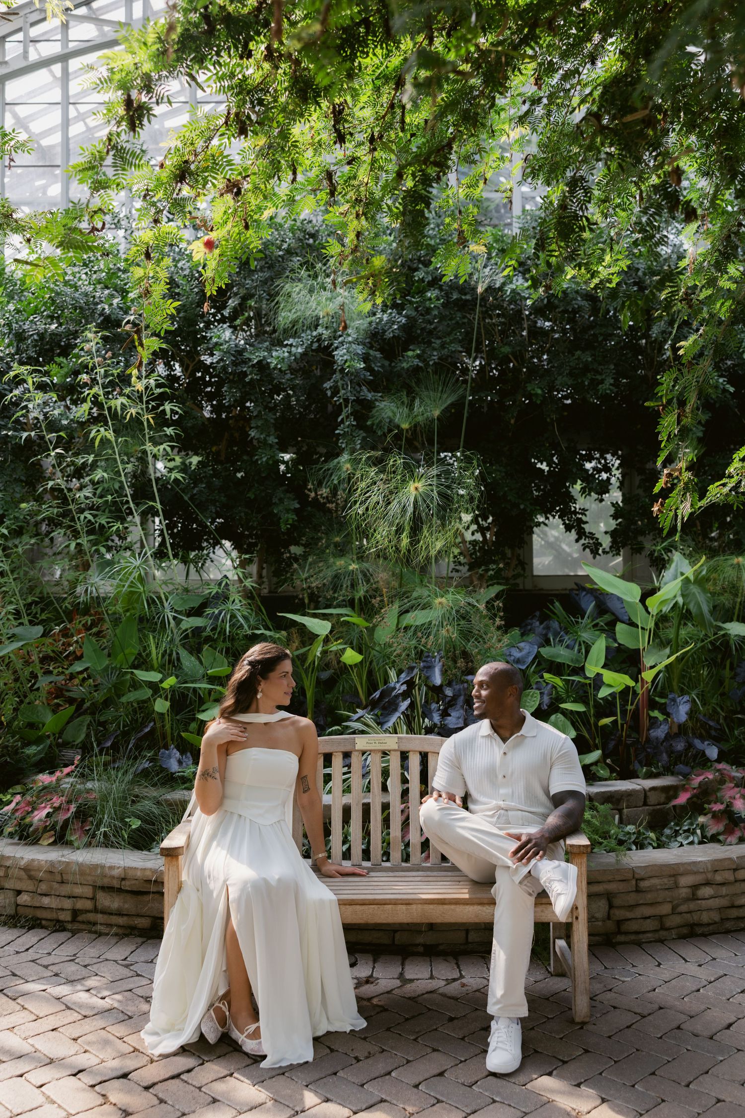 Wedding photoshoot in a botanical garden with white-clad couple sitting on benches among tropical plants.