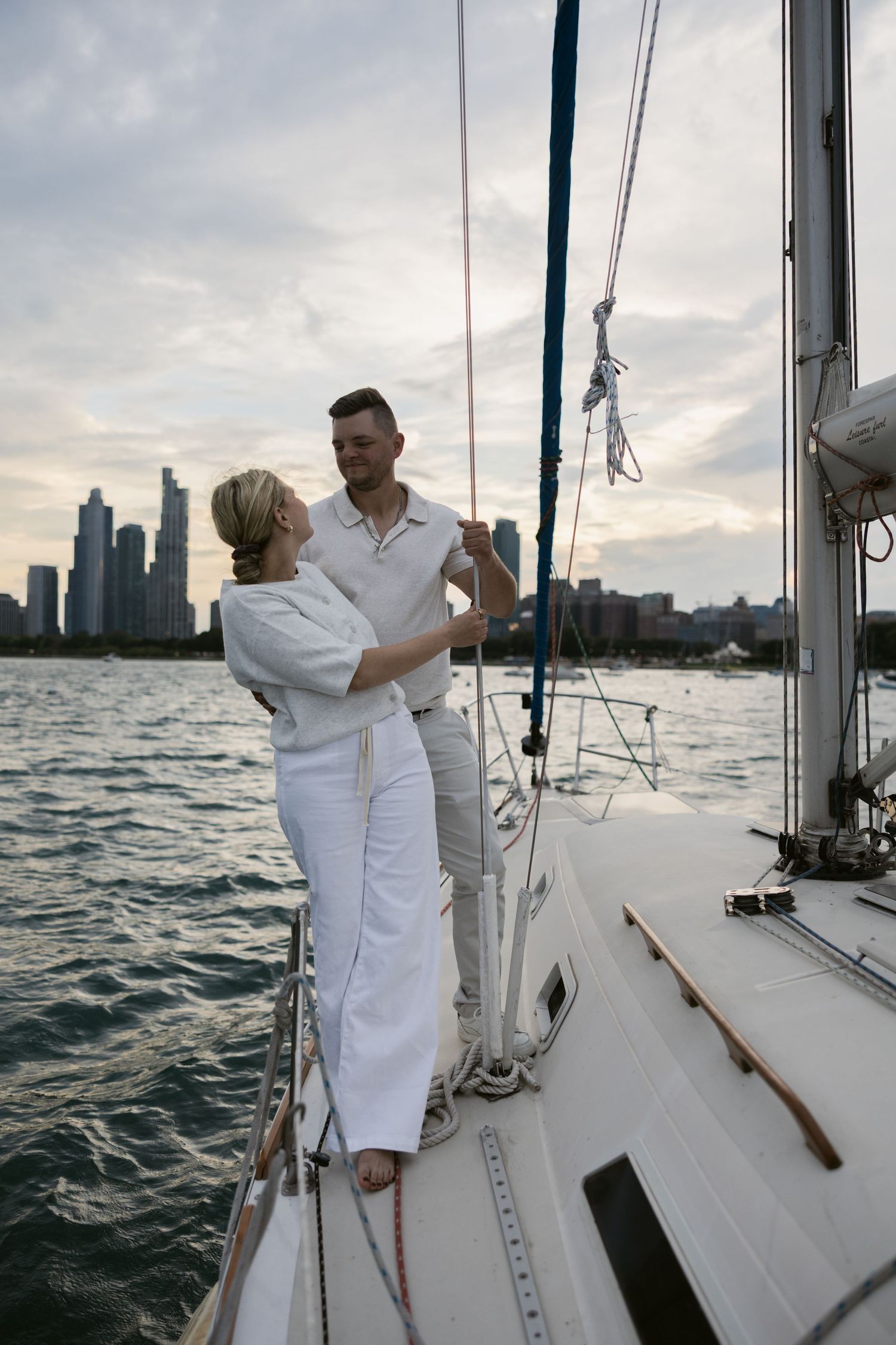 Couple in matching white outfits dancing on sailboat deck with city views.