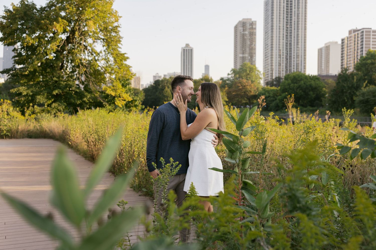 A couple shares an affectionate moment in a lush green park with tall city buildings in the background at sunset.