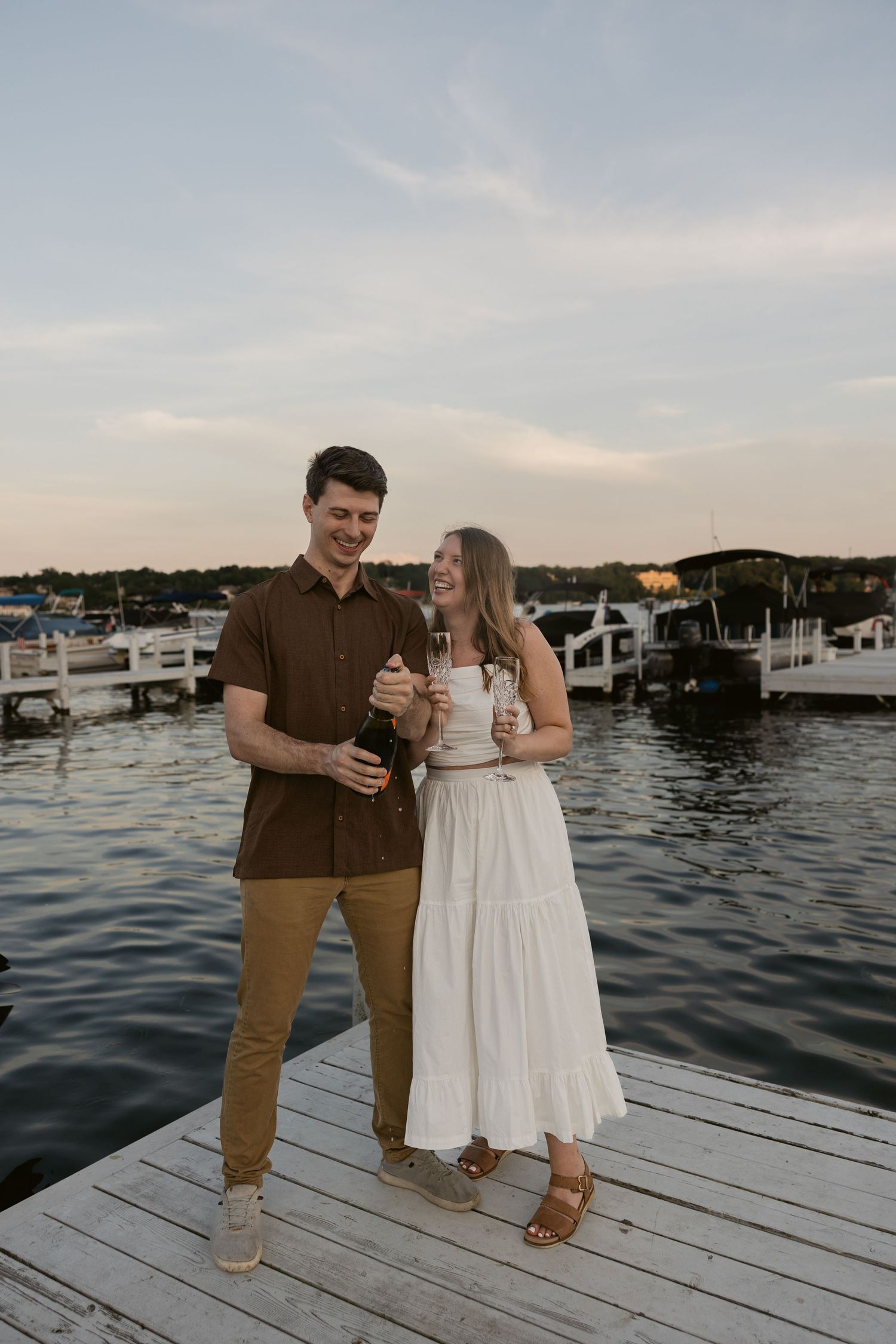 A couple shares drinks while standing on a wooden dock overlooking a marina at sunset.