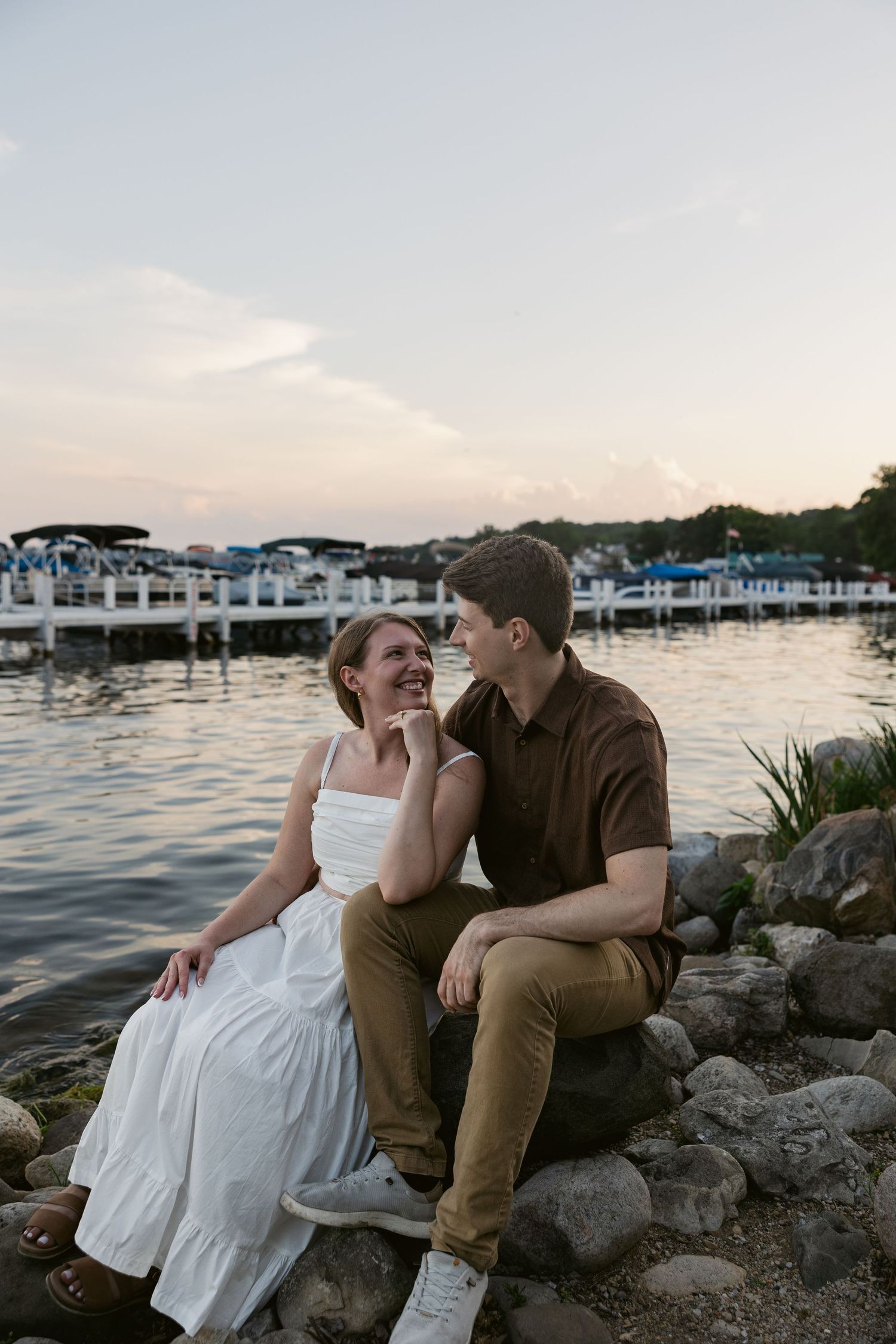 A couple sits on rocky shoreline during golden hour with waterfront townhouses in background.