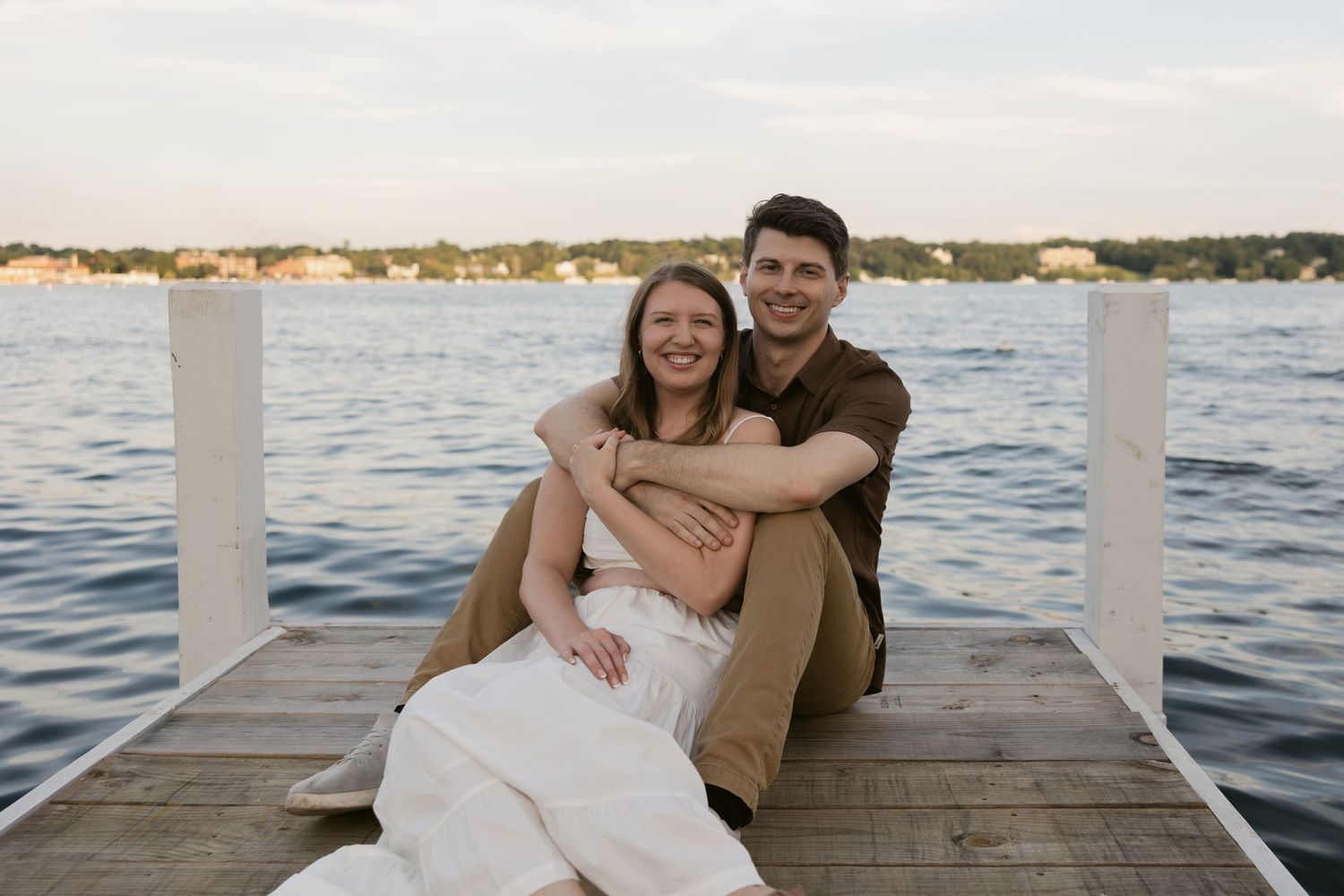 A couple sits together on a wooden dock overlooking a serene lake at sunset.