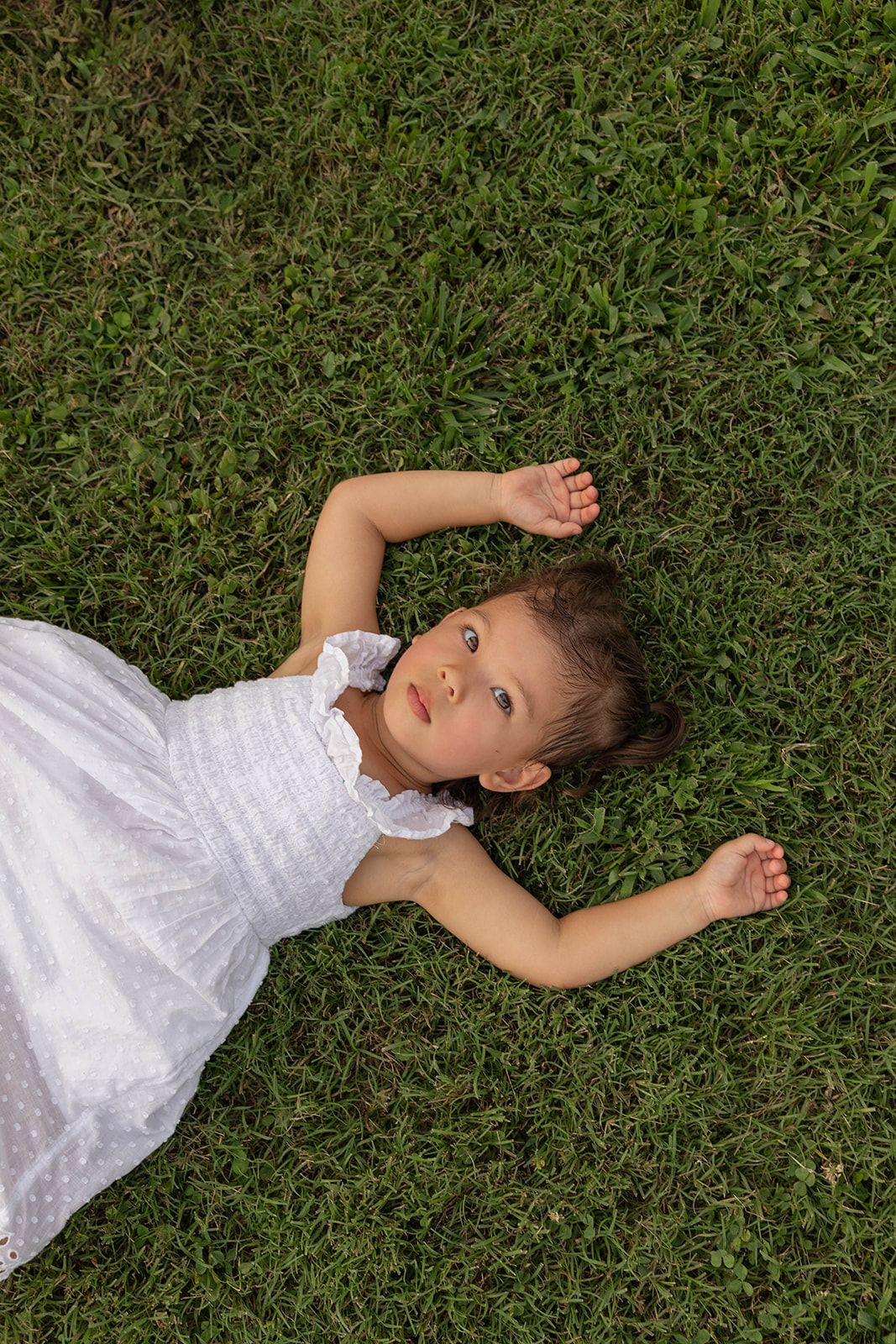 A child in a white dress lying on green grass looking up at the camera.