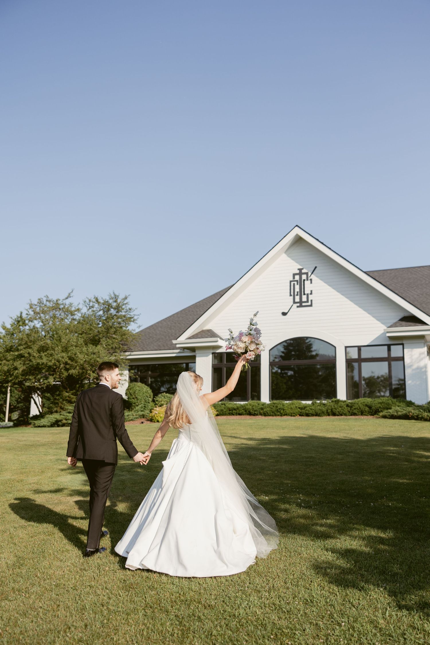 Wedding couple walking on green lawn with white buildings visible in background.