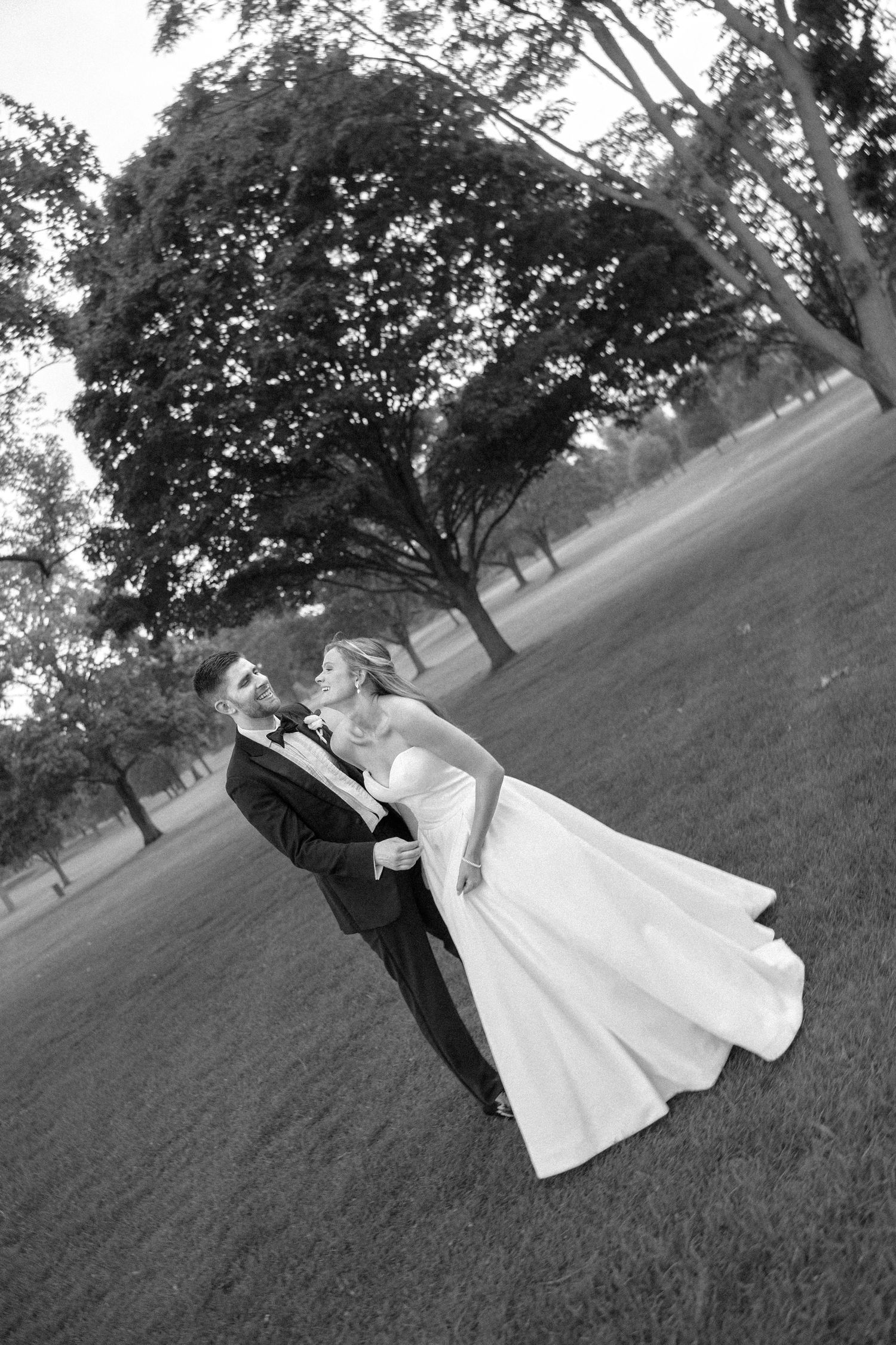 Black and white photo of a wedding couple posing playfully on a grassy field with trees.