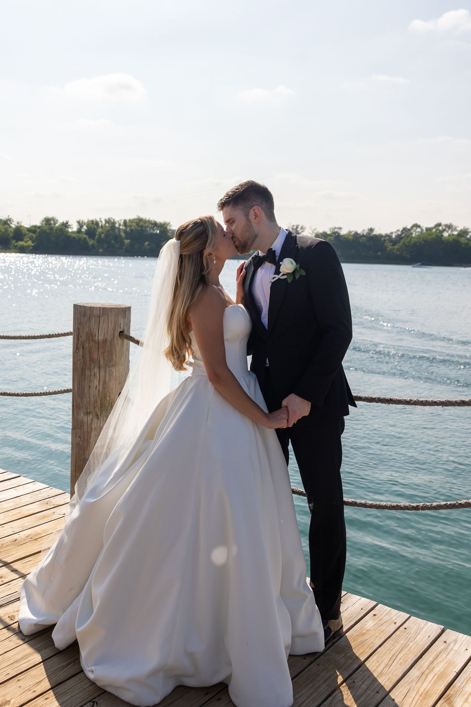 A romantic wedding couple embraces on a wooden dock overlooking a sunlit lake during golden hour.