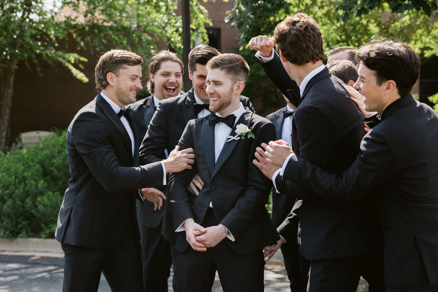 A group of groomsmen in black tuxedos laugh together outdoors.