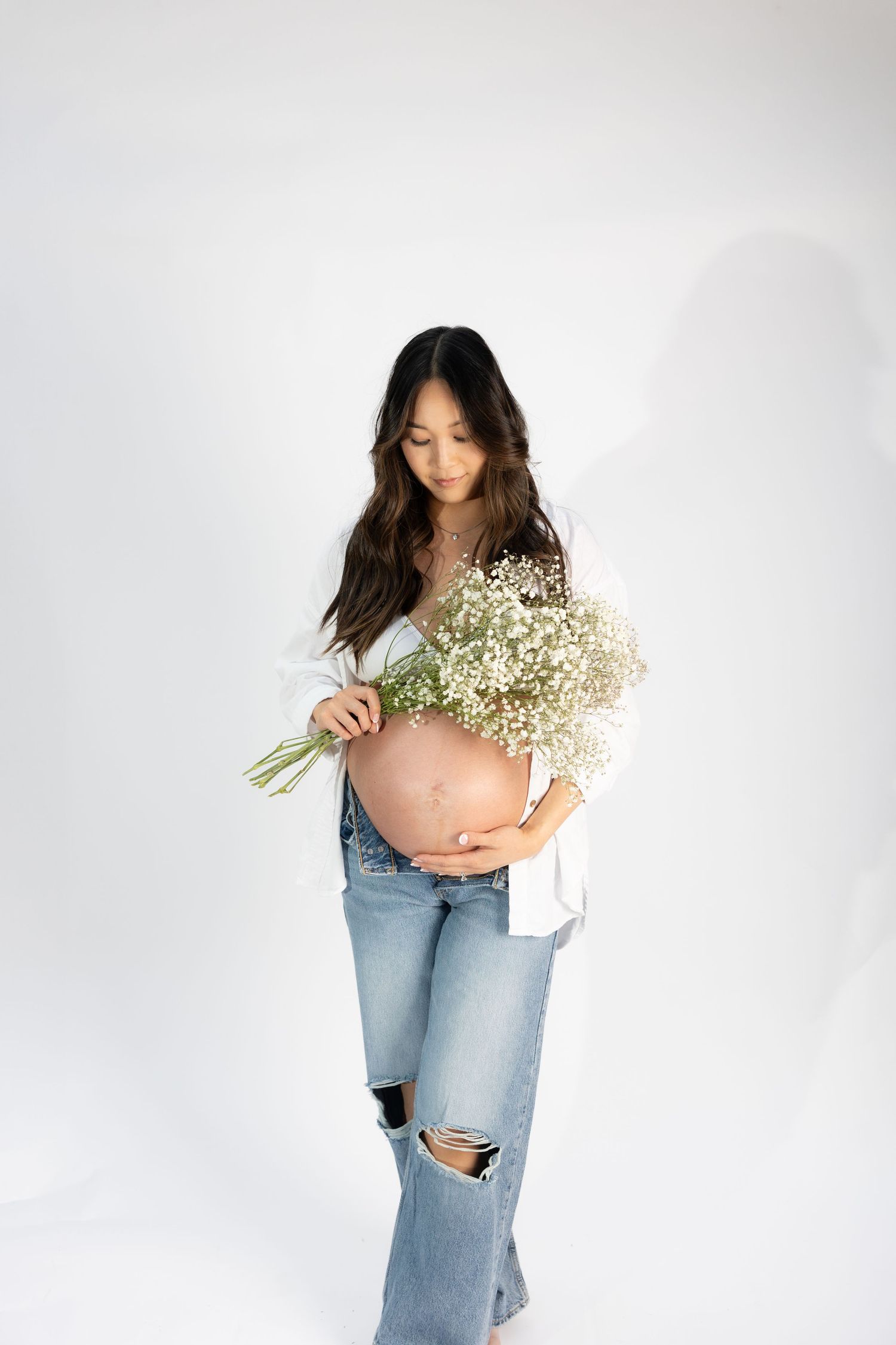 Pregnant woman in white shirt and ripped jeans holding baby's breath flowers against white backdrop.