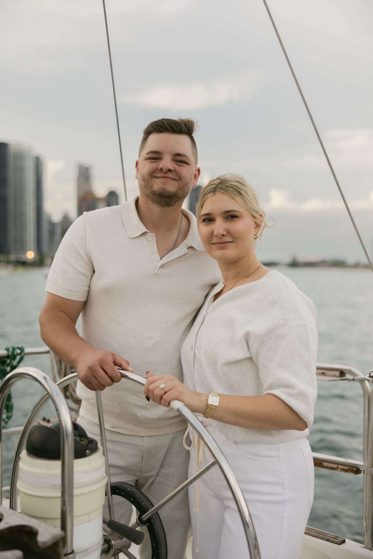 Couple in matching white outfits steering sailboat with cityscape in background.