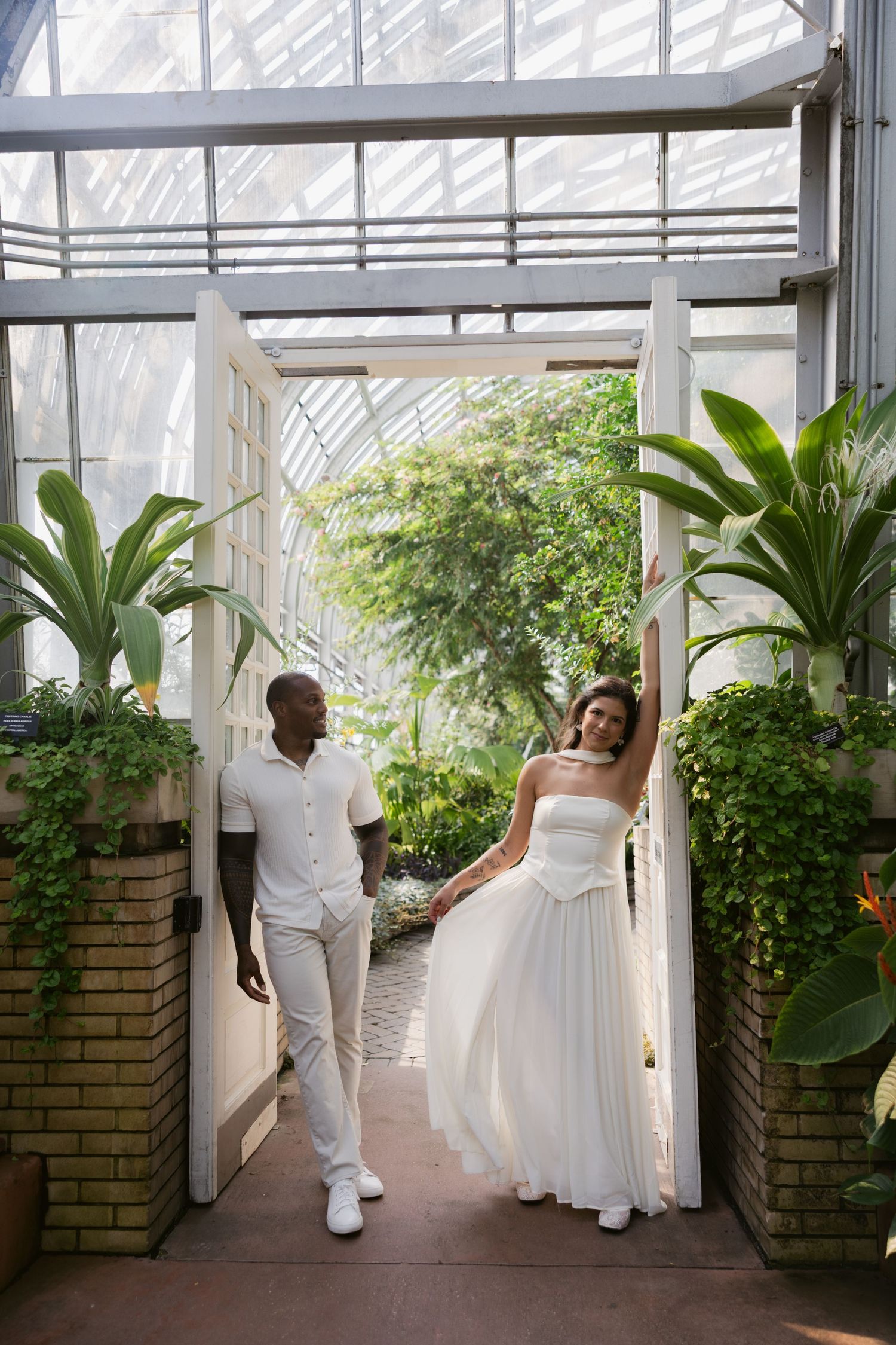 A couple in white outfits walks together through a bright greenhouse walkway surrounded by lush plants and greenery.