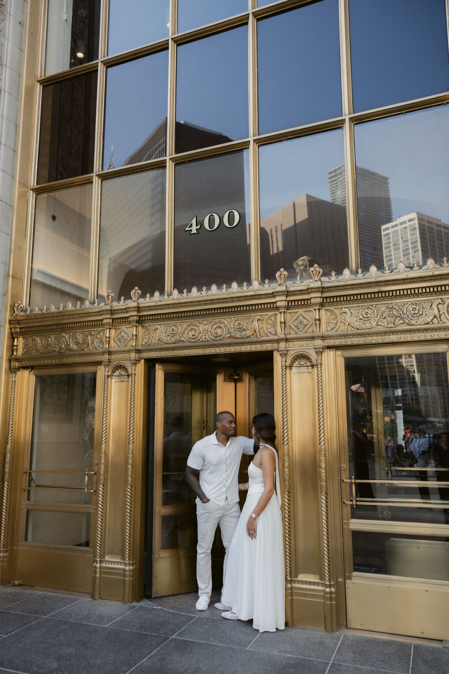 A couple stands in front of a gold-trimmed building entrance with reflective windows and architectural details.