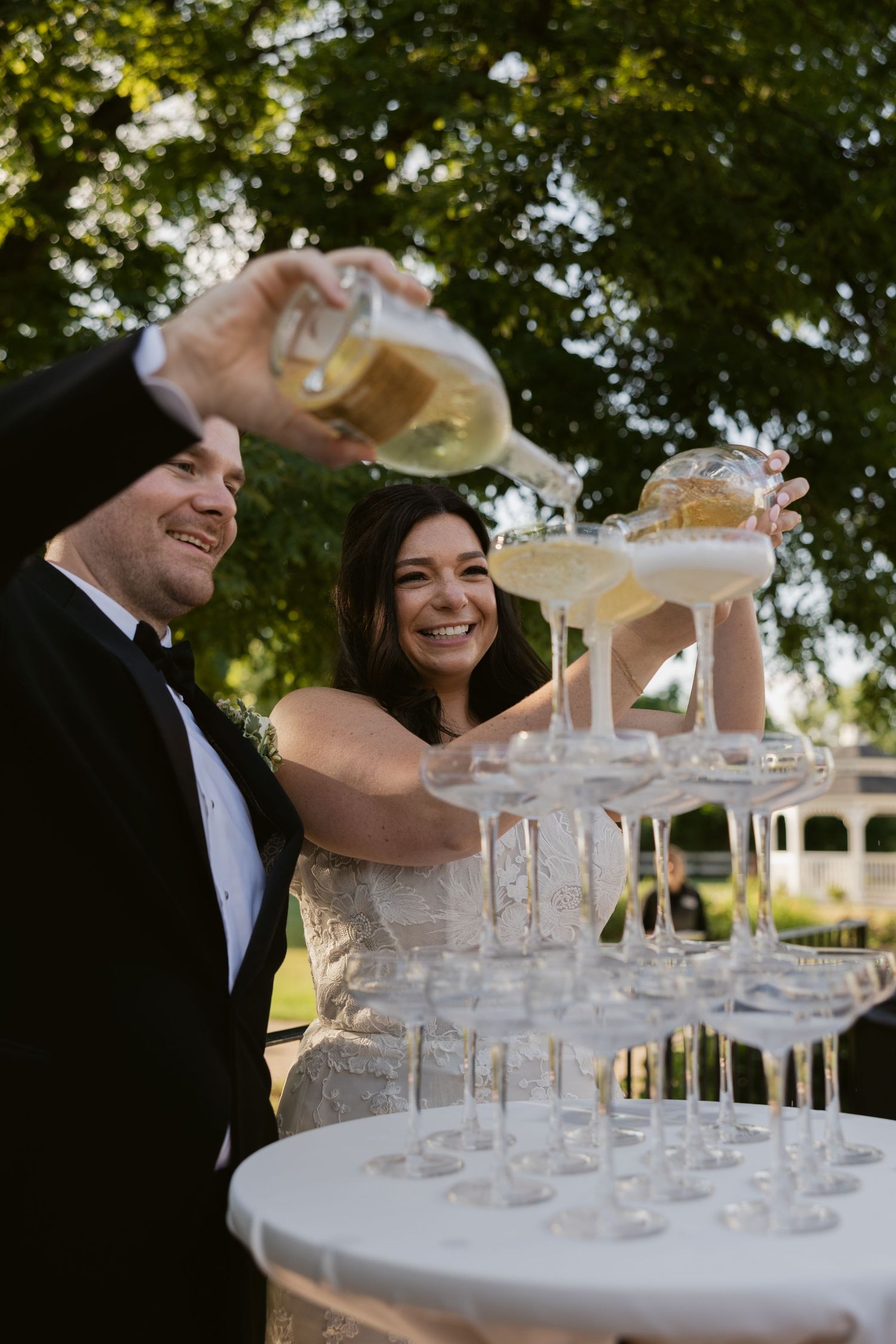 People pour champagne into a tower of stacked crystal glasses at an elegant outdoor event celebration.