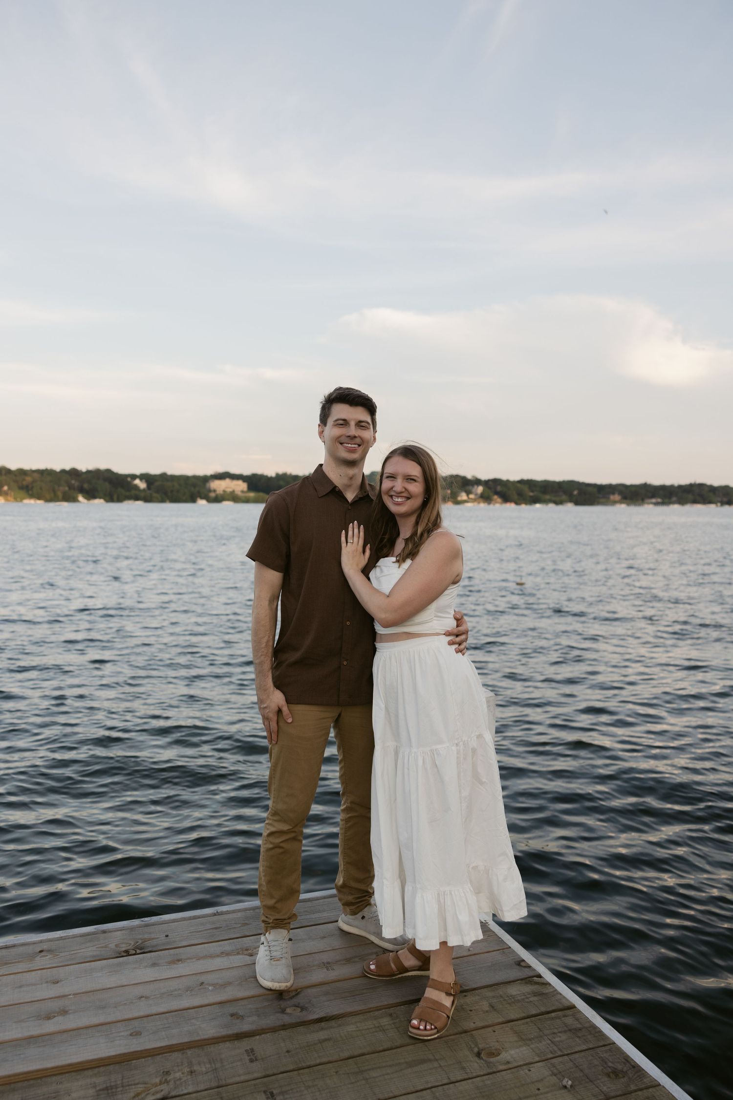 A couple stands together on a wooden dock overlooking a serene lake at dusk with blue water and distant shoreline.