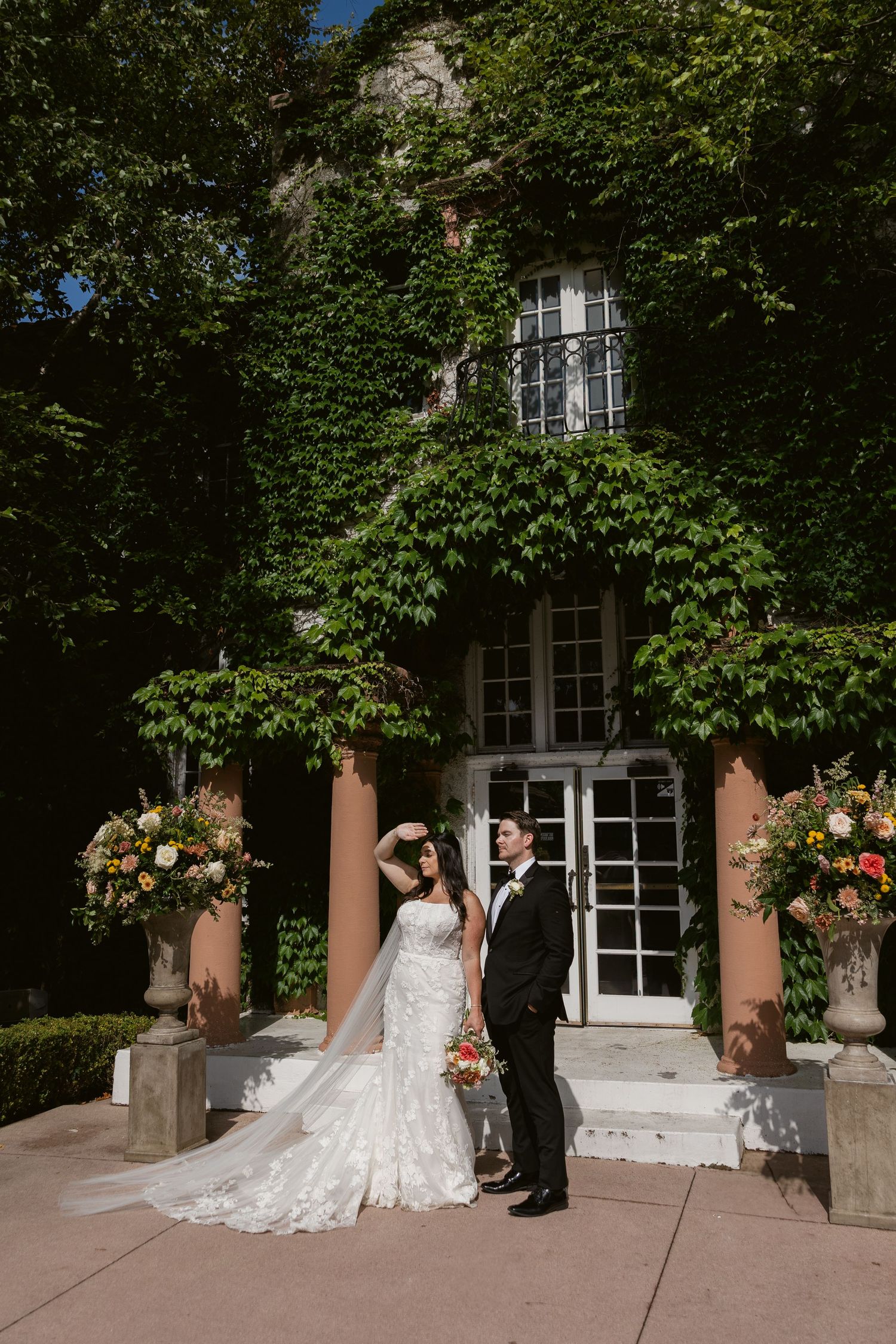 A couple pose for wedding photos outside a pink building with white trim and lush greenery.
