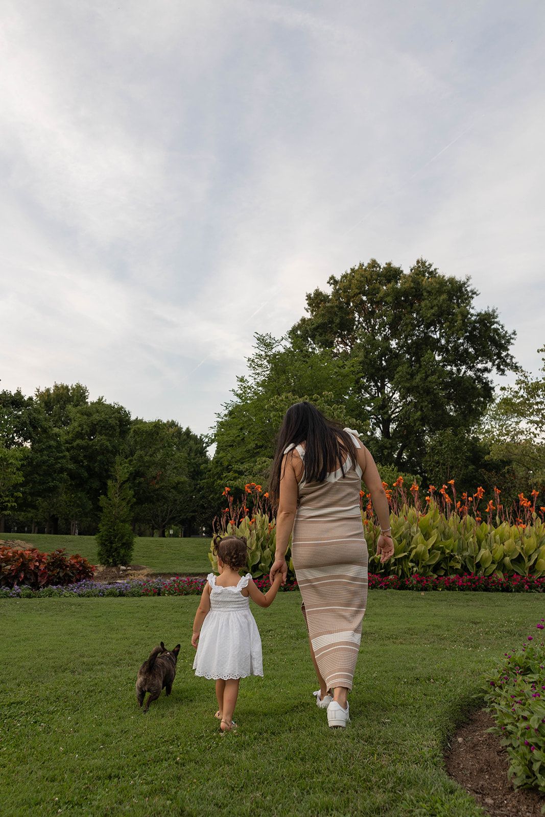 A parent and child walk hand in hand with their small dog through a garden with colorful flowers.