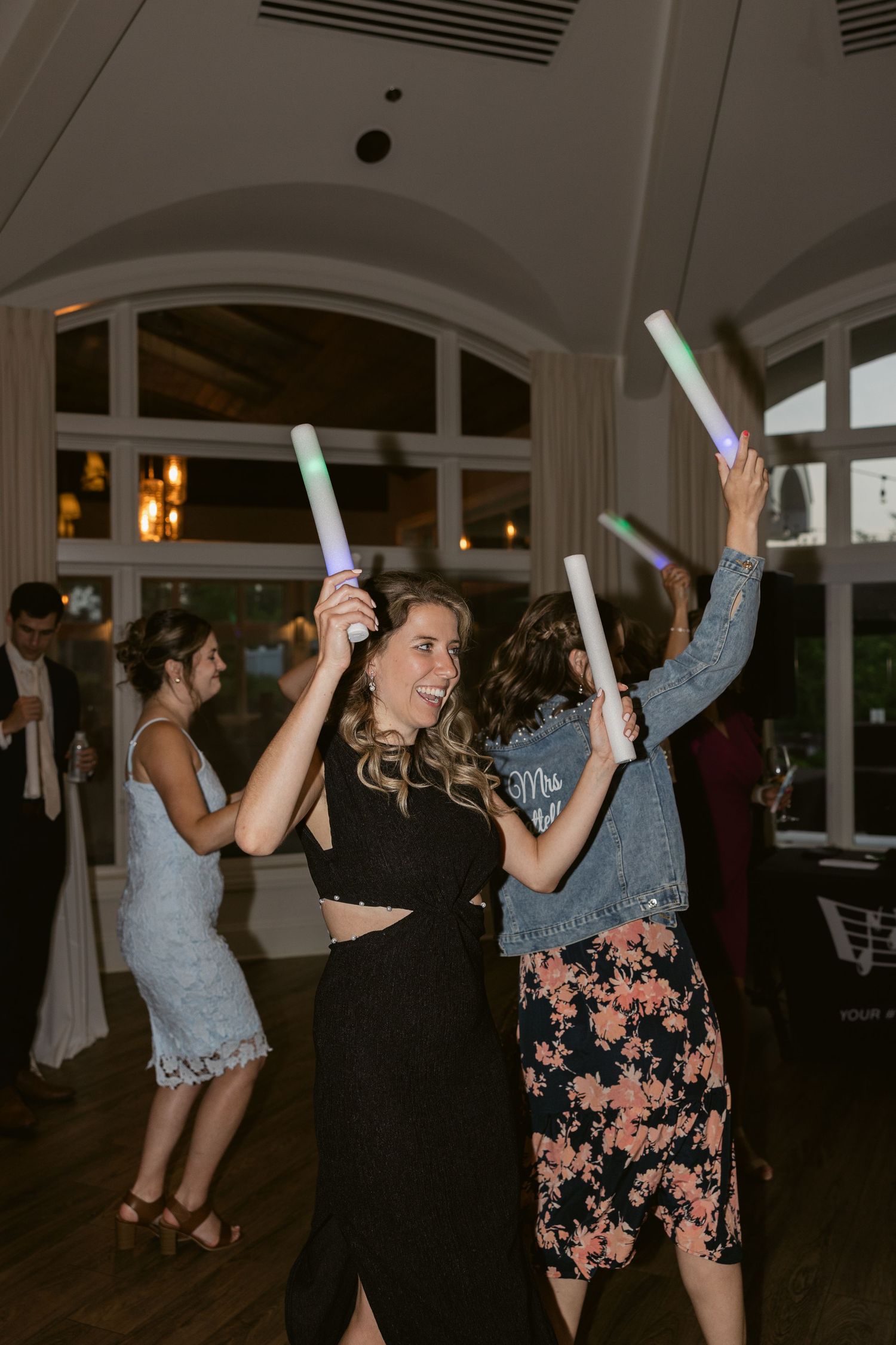 Party guests dance with glowing light sticks on a wooden dance floor at evening celebration.
